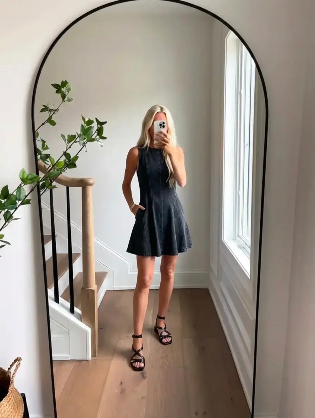 A minimalist lifestyle fashion mirror selfie in a bright modern home. A woman with long blonde hair stands in front of a large arched black-framed mirror wearing a sleeveless black denim mini dress with a fitted bodice and flared skirt, styled with black strappy sandals and delicate gold jewelry. The interior features light natural oak floors, white walls, a staircase with a light wood handrail and black metal balusters, and soft natural daylight streaming through a tall window. A leafy green plant branch adds a natural organic element. The aesthetic is effortless modern style, casual chic summer fashion, clean minimal interior, natural light lifestyle photography.

⸻

Caption

Simple, classic, and just a little bit edgy. 🖤

⸻

Hashtags

#EffortlessStyle
#BlackDress
#MinimalStyle
#CasualChic
#SummerStyle
#OOTDInspo
#ModernStyle
#TimelessFashion
#SimpleChic
#MirrorSelfieStyle
#EverydayOutfit
#StyleInspoDaily
#ChicAndSimple
#NeutralStyle
#CasualLuxury
#StyleInfluencer
#ClassicStyle
#MinimalFashion
#SummerOutfitIdeas
#StyleInspo


#LTKSpringSale #LTKgrwm #LTKdayinmylife