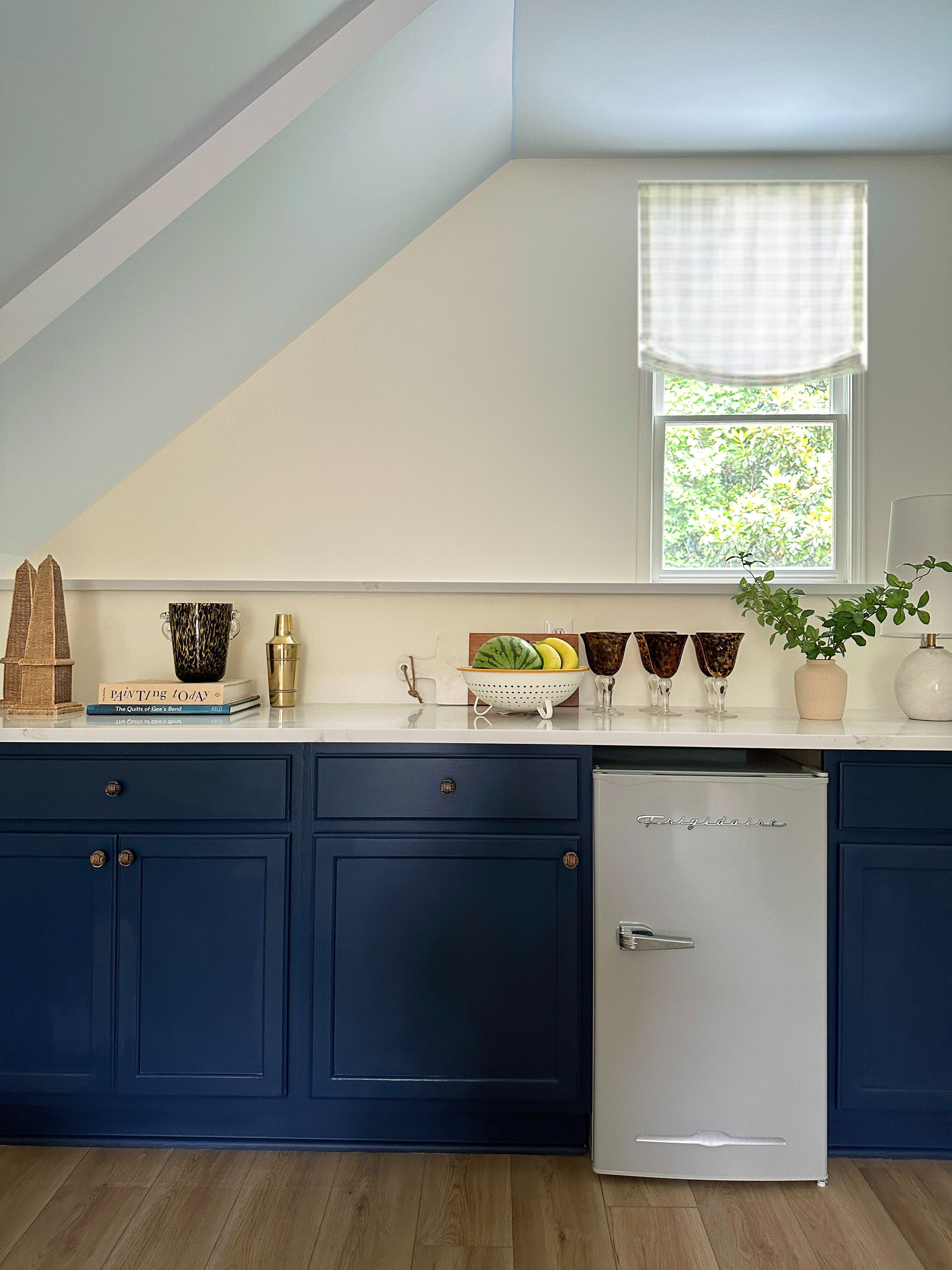 Bright and timeless kitchen design with deep navy blue lower cabinets, brushed brass hardware, and a sleek white marble countertop. The retro-inspired Frigidaire mini fridge adds charm and functionality while natural wood floors bring warmth to the space. Styled with chic glassware, fresh greenery, and a white colander filled with fruit, this kitchen corner balances modern coastal style with cozy, cottage-inspired touches. Perfect for small kitchens, guest suites, or bar areas, this setup shows how to maximize style and storage under a sloped ceiling. 

 