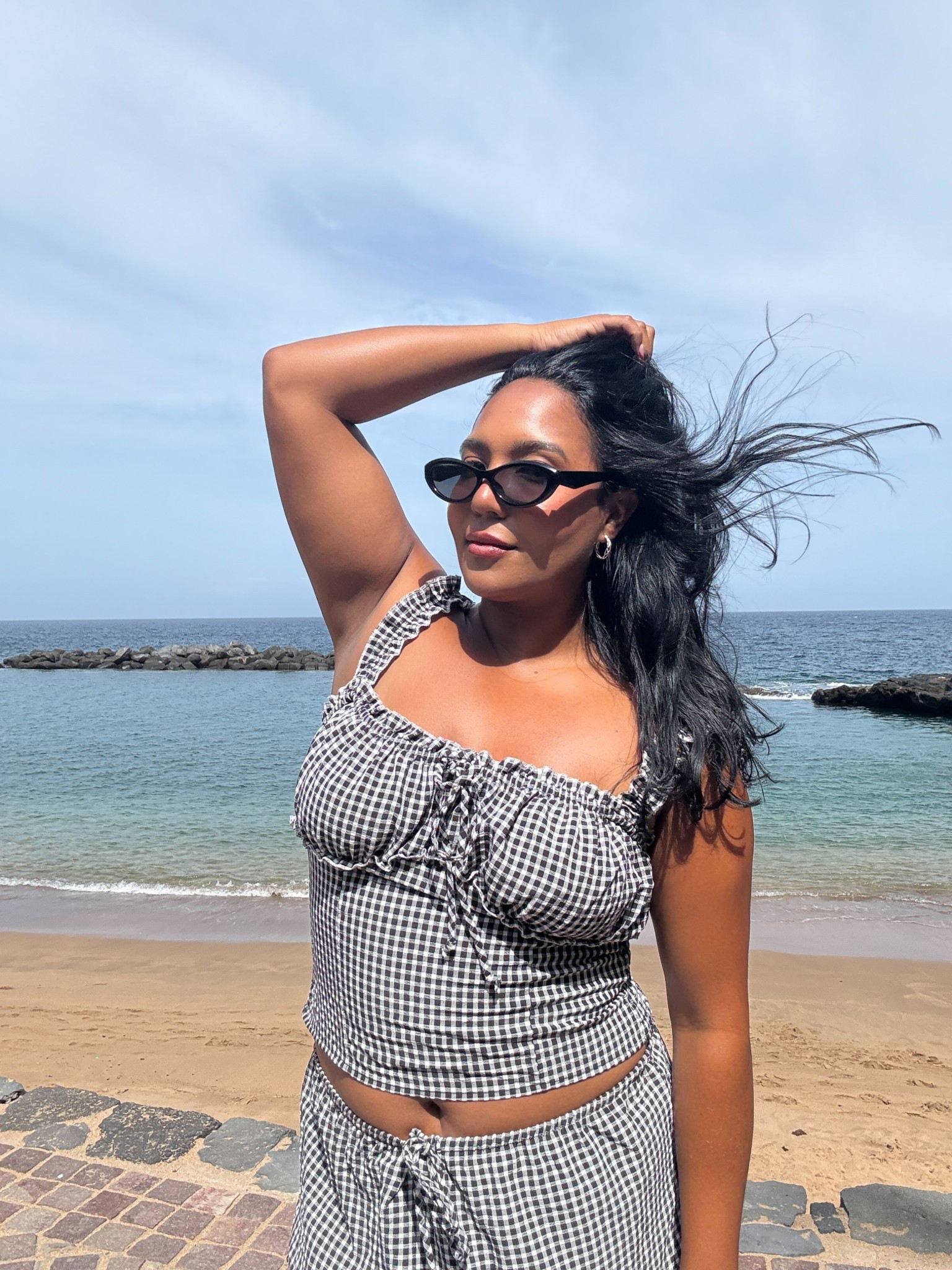 Vacation outfit vibes, but make it timeless. This gingham matching set features a bustier-style crop top with delicate ruffled straps and a high-waisted tiered skirt, perfect for a coastal grandmother aesthetic meets European summer style. The black and white check print keeps it classic yet playful, while cat-eye sunglasses and soft beach waves complete the effortless chic look. Ideal for midsize-friendly resort wear, travel outfit inspo, or a dreamy Mediterranean-inspired getaway.