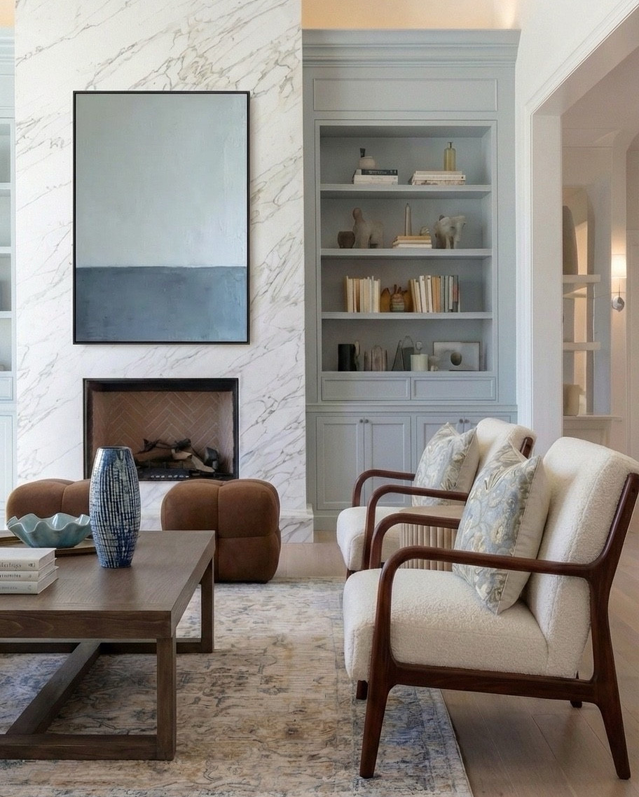 Shop this look:
• Exposed wood ceiling beams
• Blue floral wallpaper (ceiling + vaults)
• Marble fireplace surround
• Gas fireplace insert
• Framed neutral landscape artwork
• Built-in bookcases with lower cabinetry (soft blue-gray)
• Styled books + neutral ceramic decor
• Cream upholstered sofa
• Blue chunky knit throw blanket
• Blue and neutral accent pillows (patterned + solid)
• Upholstered accent chairs with wood frames
• Brown leather ottomans
• Rectangular wood coffee table
• Blue ceramic decorative vase
• Blue decorative bowl
• Stacked coffee-table books
• Light blue patterned area rug
• Small round wood side table
• Table lamp with white shade
• Floor-to-ceiling windows with sheer white curtains

#LTKHome #LTKSaleAlert #LTKSpringSale