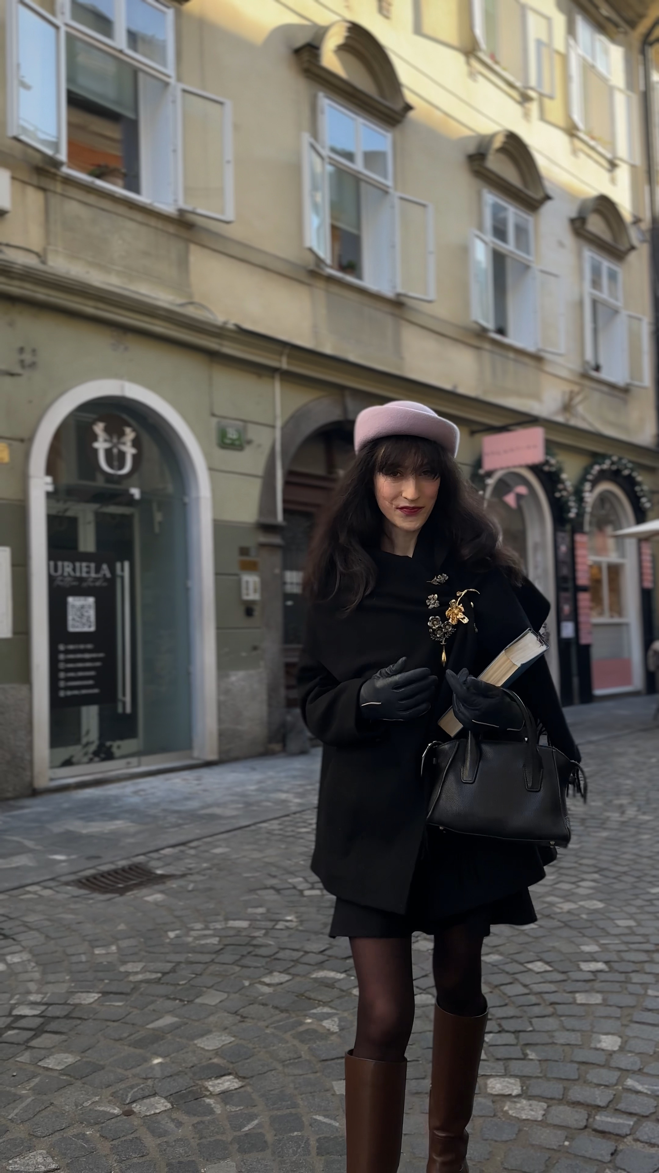 Winter street style 2026, black wool coat outfit, vintage gold brooch styling, pink hat winter look, brown riding boots outfit, European cobblestone streets, Parisian winter aesthetic, old money winter style, black and brown combination, leather gloves winter outfit, structured handbag outfit, carrying books aesthetic, sophisticated winter layers, elegant cold weather fashion, Europe street style 

 #LTKdayinmylife #LTKootd #LTKTravel