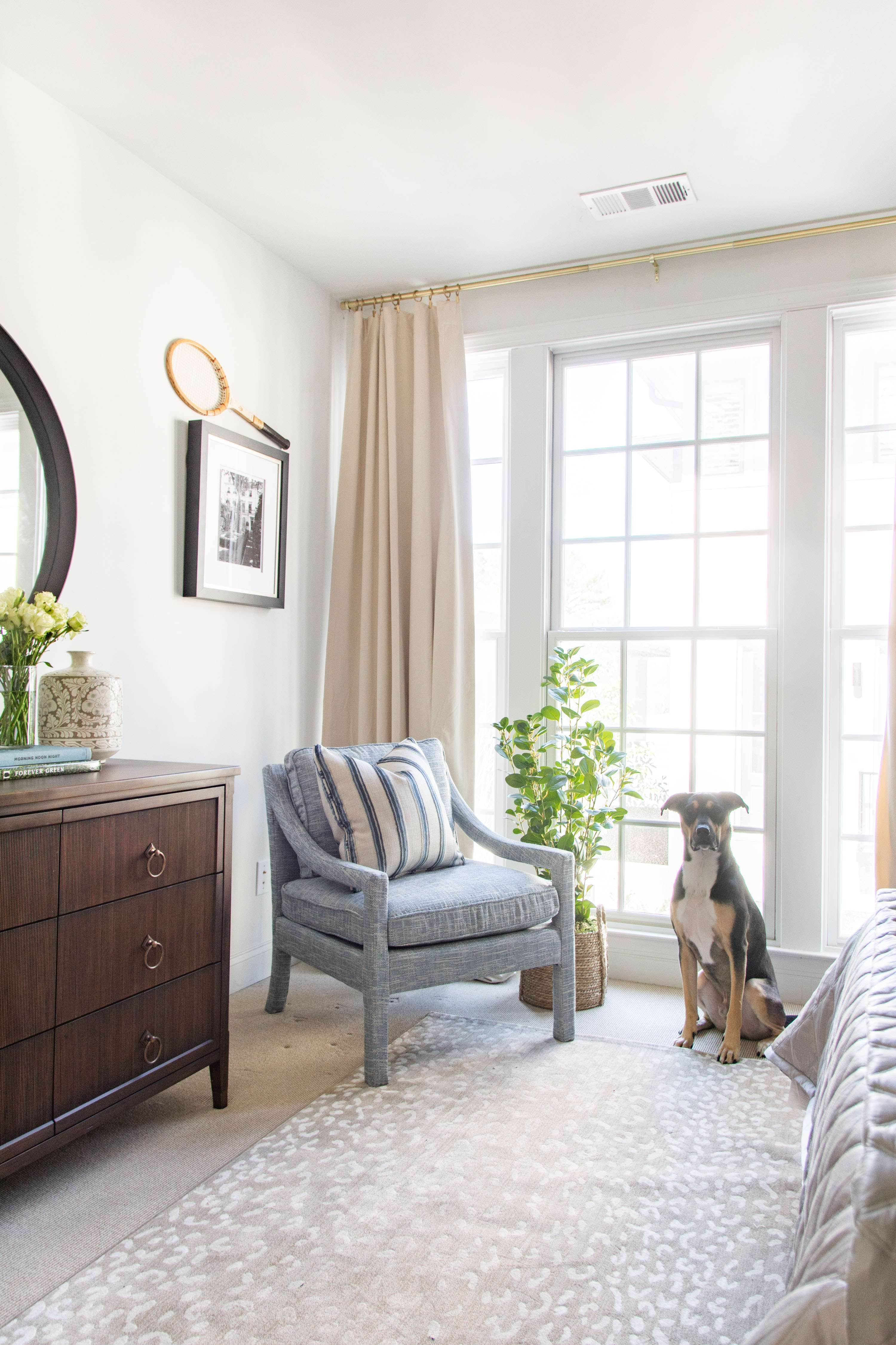Bright and airy bedroom corner design featuring floor-to-ceiling windows with neutral curtains, a modern gray accent chair with striped pillows, and a warm wood dresser styled with fresh flowers and decor accents. The cozy space is layered with a light patterned rug, natural textures, and greenery for a fresh, inviting feel. Perfect inspiration for transitional home decor, neutral bedroom design, and modern classic interiors with a touch of coastal charm. 

 