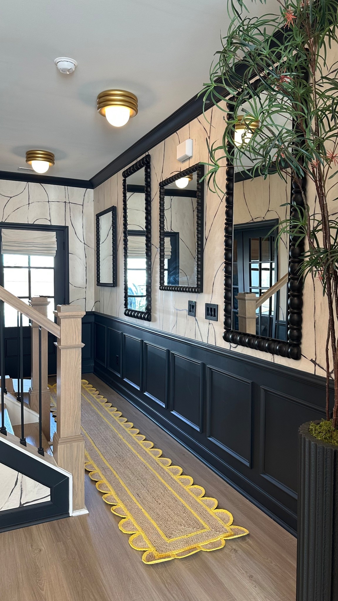 Entryway makeover with black wainscoting mirrors, faux tree, gold flush mount lighting and whimsical scalloped yellow jute runner rug 