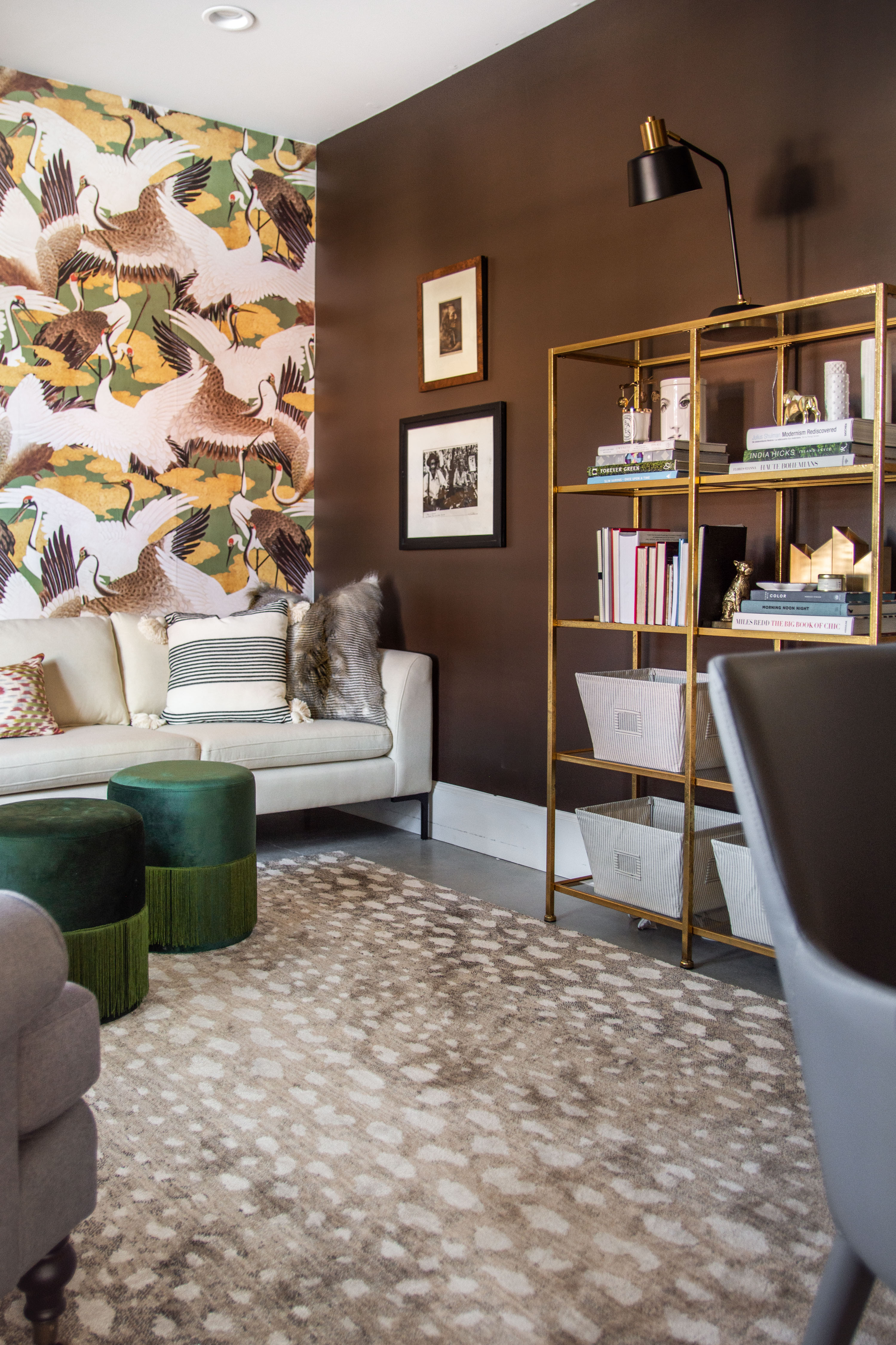 Modern living room design inspiration featuring a bold statement wall with crane-patterned wallpaper, paired with a sleek white sofa and layered textured pillows. The space combines luxe elements like emerald green velvet ottomans with fringe, a warm chocolate brown accent wall, and a gold open shelving unit styled with books and decor. A neutral patterned rug grounds the space while mixing contemporary and eclectic design details. Perfect for anyone searching for living room makeover ideas, wallpaper accent wall inspiration, or chic home decor styling. 

 