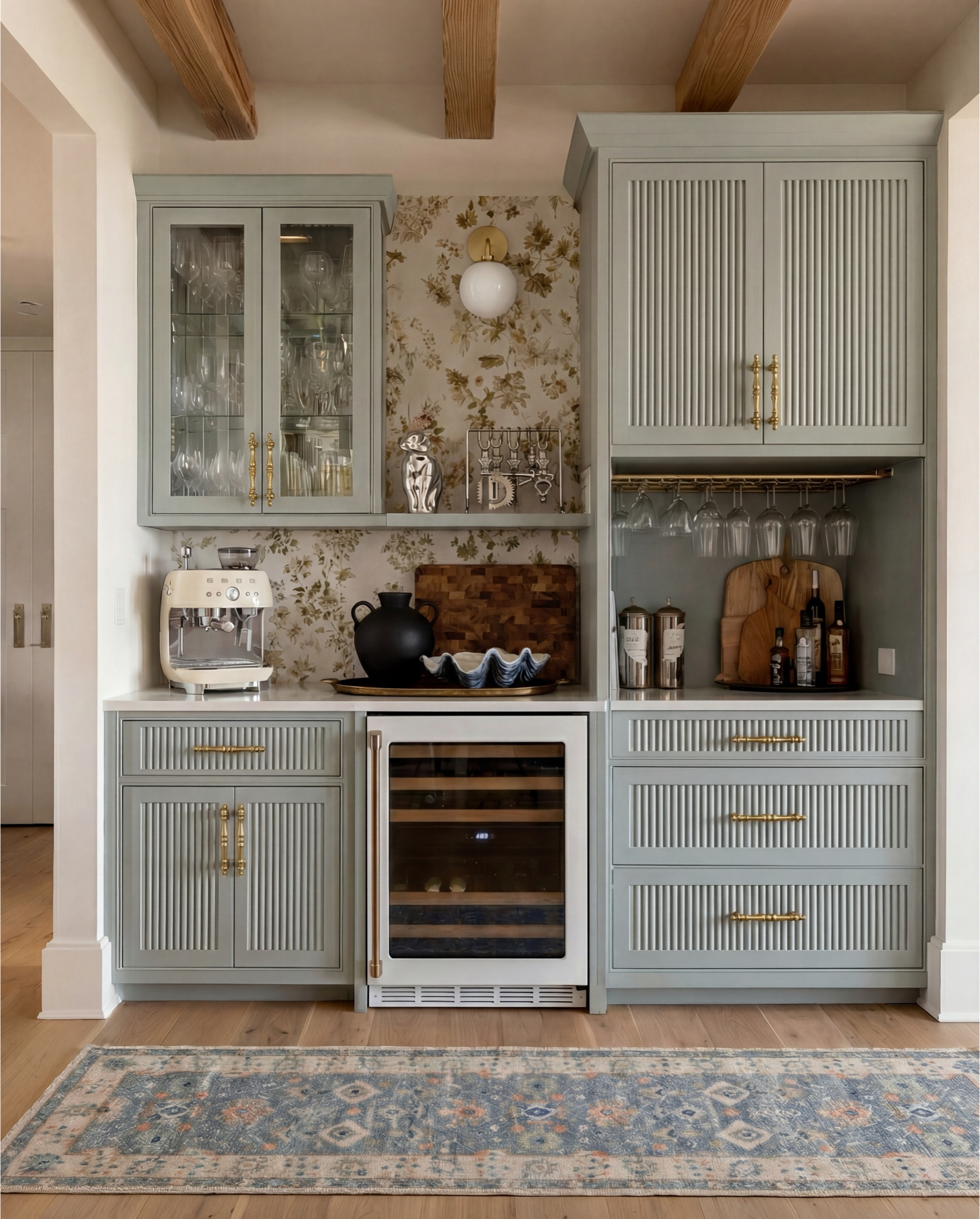 This cabinet bar area feels refined, warm, and quietly elevated. Soft sage cabinetry with fluted detailing creates a tailored, custom look, while aged brass hardware and glass-front uppers add a classic, collected feel. The floral wallpaper backdrop softens the space with subtle romance, balanced by natural wood accents and warm neutral tones that keep everything grounded and inviting.

The styling feels layered yet intentional—vintage-inspired textures, sculptural ceramics, and functional bar essentials that turn a simple cabinet wall into a cozy entertaining moment.

Shop the Look:
• Aged brass cabinet pulls and knobs
• White built-in wine fridge
• Cream Smeg espresso machine (retro style)
• Wood cutting boards (mixed warm wood tones)
• Matte black ceramic pitcher
• Scalloped ceramic serving bowl (soft neutral)
• Under-cabinet stemware rack with wine glasses
• Floral neutral wallpaper (soft beige + gold tones)
• White globe brass wall sconce
• Vintage-style blue and beige runner rug
• Floating display shelf with decorative accents
•Drink martini shakers 
Black vase
Brass tray
• Neutral stone or quartz countertop

#LTKSaleAlert #LTKHome