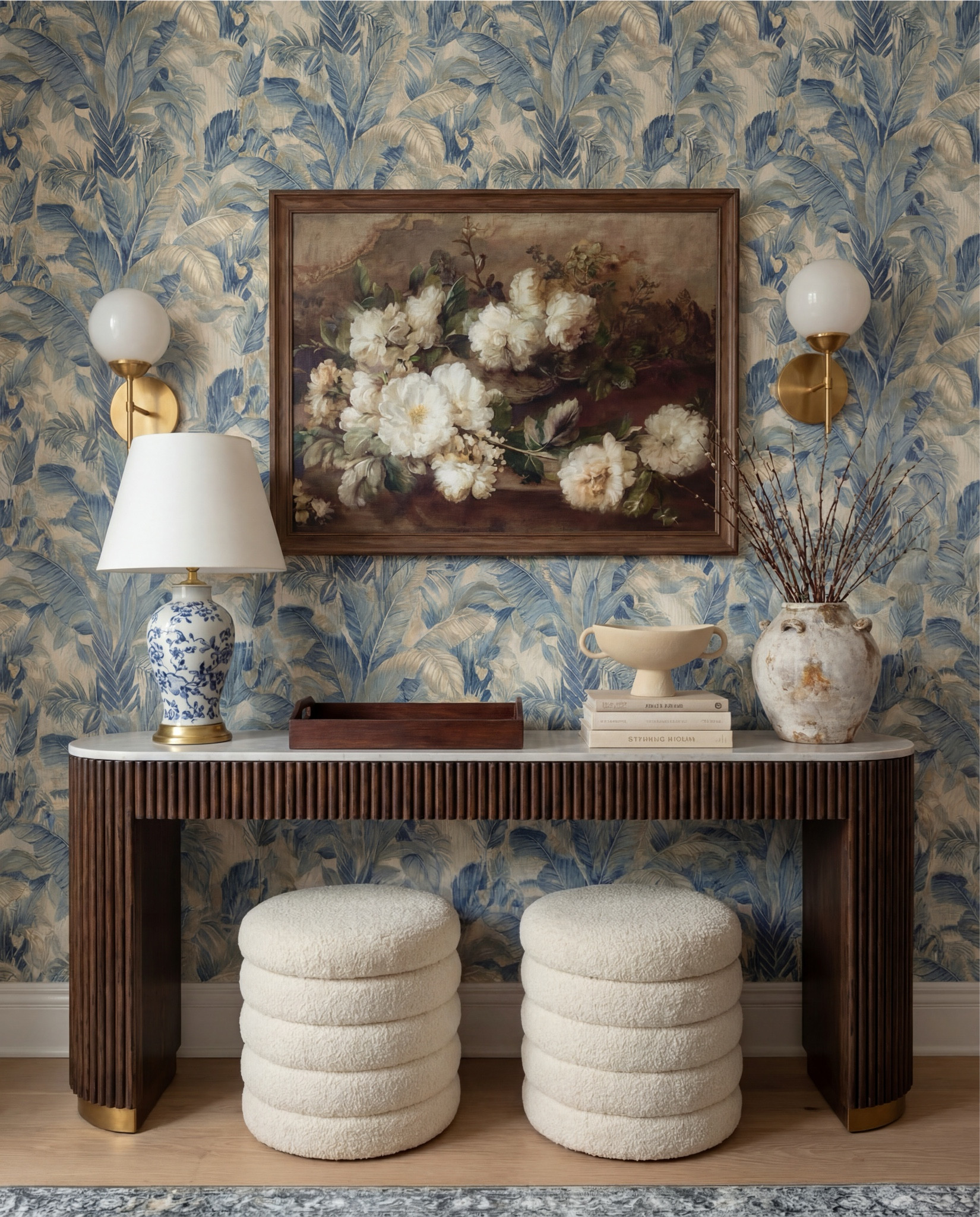 This foyer feels classic, layered, and quietly elegant—the kind of entry that instantly signals a thoughtfully designed home. The blue botanical wallpaper brings softness and movement, while warm wood tones and brass accents ground the space and keep it from feeling too formal. It’s traditional at first glance, but the textures and silhouettes make it feel fresh and livable.

The styling is balanced and intentional—symmetry at the wall, contrast in materials, and sculptural pieces that feel collected over time rather than styled all at once.

Shop the Look:
• Blue botanical wallpaper (leaf motif, muted navy + beige)
• Solid wood console table (dark walnut finish)
• Fluted wood console table legs
• White marble console tabletop
• Framed vintage floral wall art (dark wood frame)
• Brass wall sconces with white globe shades
• Blue and white ceramic table lamp
• White fabric lamp shade (tapered)
• Wooden decorative tray (rectangular)
• Stacked neutral coffee table books
• Sculptural ceramic bowl (cream stoneware)
• Distressed ceramic vase (antique white)
• Dried branches or twigs arrangement
• Bouclé ottomans (ivory, round)
• Light wood flooring
• White baseboards and trim

#LTKHome #LTKSaleAlert #LTKFindsUnder100
