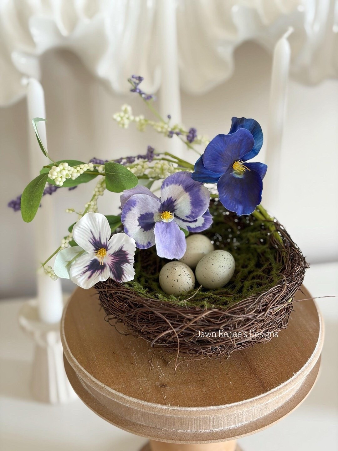 Add a sweet touch of spring to your décor with this charming birds nest sourced by DawnReneesDesigns. Made with natural twig and moss, the nest features three faux speckled eggs surrounded by delicate pansy blooms, tiny berries, and greenery for a fresh garden-inspired look. Measuring about 6” wide with greenery extending to roughly 9”, it’s perfect for styling in wreaths, centerpieces, or seasonal displays that celebrate the beauty of spring. #springdecor #easterdecor #nestdecor #farmhousestyle #ltkhome #seasonaldecor #homedecorfinds 

#LTKSeasonal #LTKHome