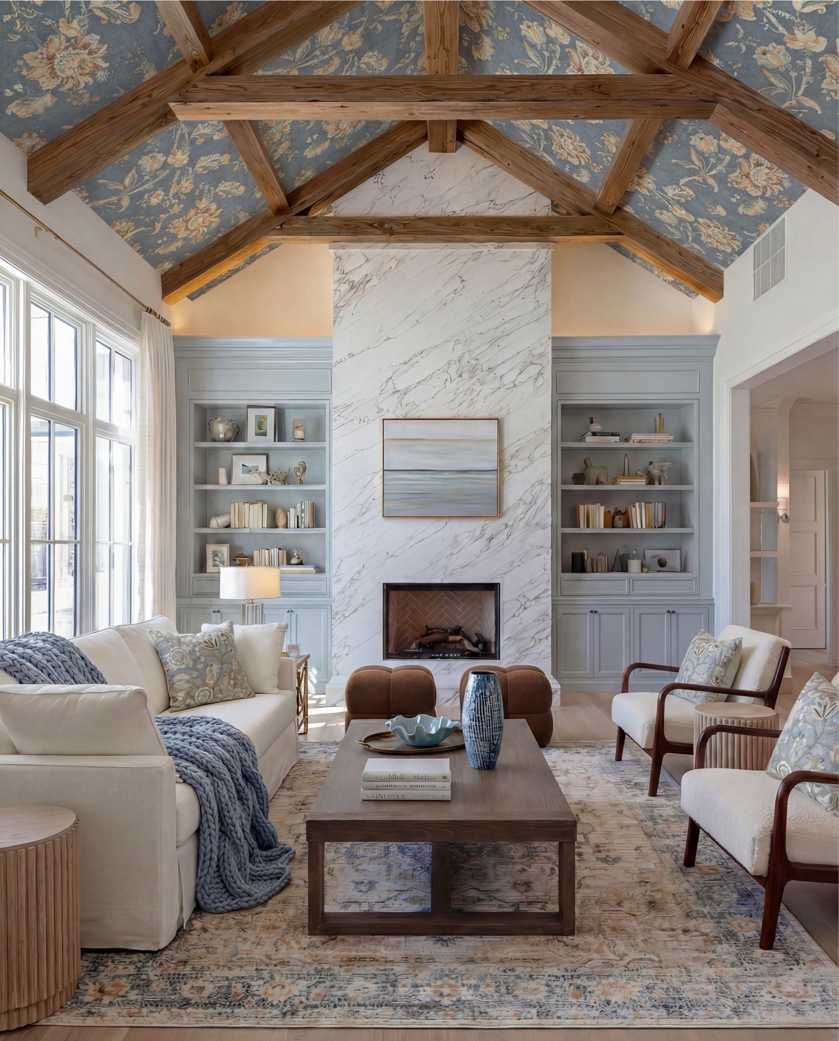 Soft blues, warm woods, and architectural balance—this living room is a textbook traditional-modern moment. The floral ceiling wallpaper and exposed beams add drama, while tailored upholstery and clean-lined furniture keep it grounded and livable.

Shop this look:
• Exposed wood ceiling beams
• Blue floral wallpaper (ceiling + vaults)
• Marble fireplace surround
• Gas fireplace insert
• Framed neutral landscape artwork
• Built-in bookcases with lower cabinetry (soft blue-gray)
• Styled books + neutral ceramic decor
• Cream upholstered sofa
• Blue chunky knit throw blanket
• Blue and neutral accent pillows (patterned + solid)
• Upholstered accent chairs with wood frames
• Brown leather ottomans
• Rectangular wood coffee table
• Blue ceramic decorative vase
• Blue decorative bowl
• Stacked coffee-table books
• Light blue patterned area rug
• Small round wood side table
• Table lamp with white shade
• Floor-to-ceiling windows with sheer white curtains

#LTKHome #LTKSaleAlert #LTKSpringSale