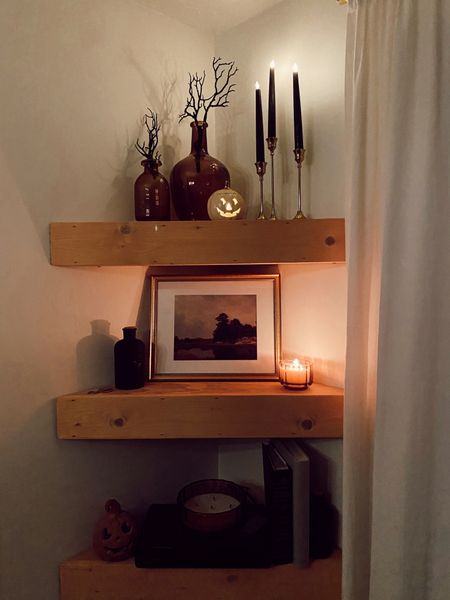 I love decorating these 3 corner light wood shelves for the seasons. Here is how I decorated them for Halloween! On the top shelf their are two amber glass bottle vases with black glittery branches in them. 3 black tapered faux candle sticks in gold candle stick holders. A small white jack o lantern. On the middle shelf there is a framed piece of moody artwork, a lit fall candle in an amber glass container, with a spells bottle on the other side. On the bottom shelf there is some photo albums and coffee table books, a hidden cable box and one of my DIY terracotta pumpkins that are pottery barn dupe!

#LTKHalloween #LTKSeasonal #LTKhome