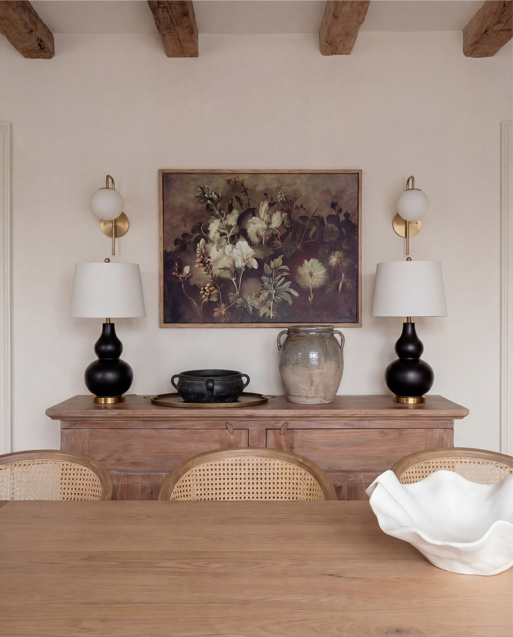 Shop the Look:
• Solid wood dining table (natural oak tone)
• Cane-back dining chairs with upholstered seats
• Upholstered accent dining chair (cream boucle)
• Vintage-style area rug (neutral, muted pattern)
• Exposed wood ceiling beams
• White globe pendant light with brass stem
• Brass wall sconces with globe shades
• Wood sideboard/buffet cabinet
• Black table lamp with white shade
• Ceramic table lamp (neutral glaze)
• Framed abstract artwork
• Glass-front wood hutch cabinet
• White sculptural ceramic bowl (table centerpiece)
• Potted olive tree (faux or real)
• Light wood flooring
• Large windows with simple trim


#LTKHome #LTKSaleAlert