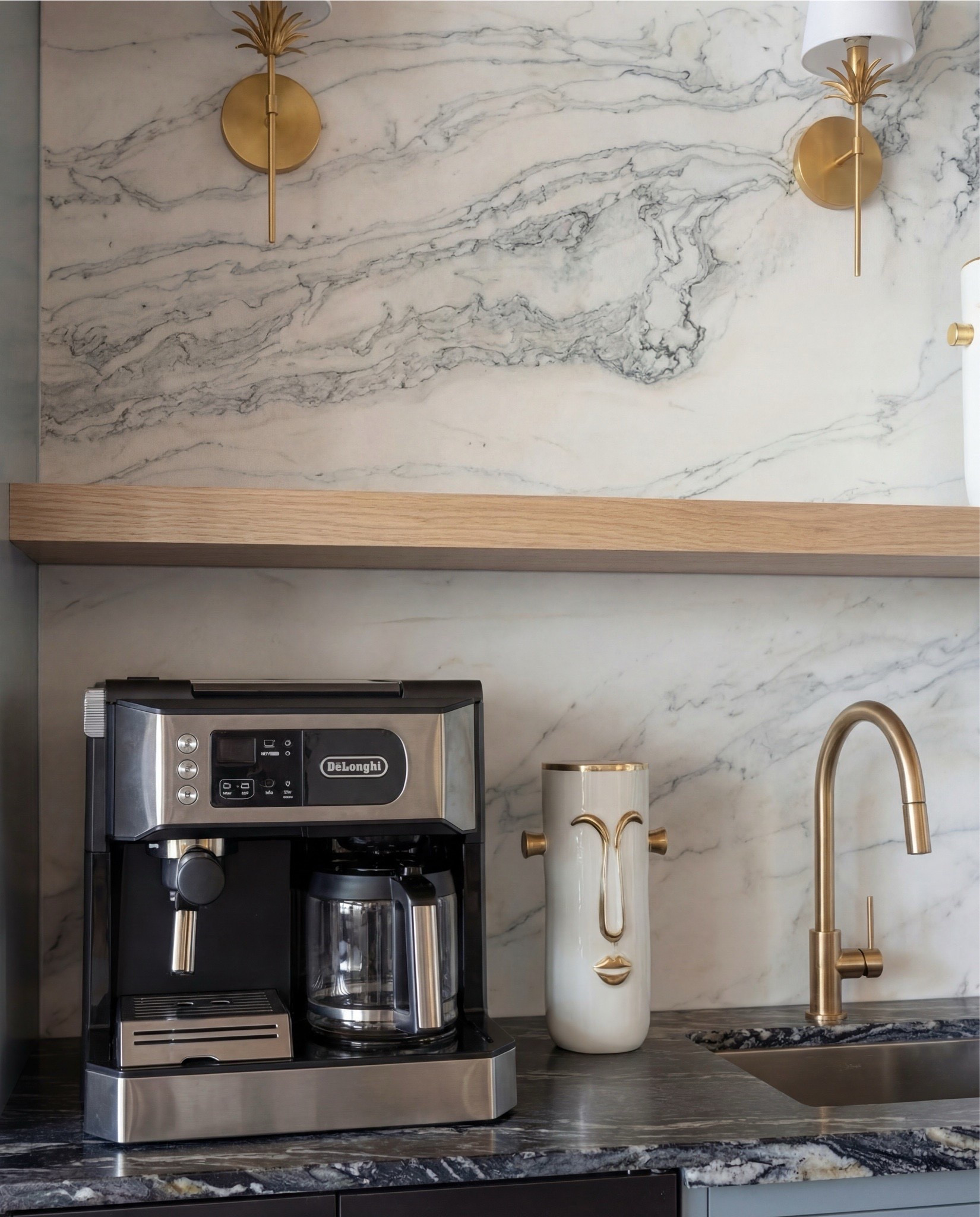 This cabinet moment feels polished, architectural, and effortlessly elevated. The soft blue cabinetry brings a calm, tailored presence, while the dramatic marble backsplash adds movement and contrast. Warm brass details—from the hardware to the sconces and faucet—layer in a quiet luxury that feels timeless rather than trendy. Dark wood ceiling beams and a simple oak shelf ground the space and keep it from feeling too formal.

The styling is minimal but thoughtful—functional pieces double as decor, letting the materials and finishes take center stage.

Shop the Look:
• Soft blue paneled cabinetry
• Brass cabinet pulls + drawer handles
• Fluted glass upper cabinet doors
• Marble slab backsplash (gray veining)
• Dark marble countertops
• Brass wall sconces with white shades
• Built-in beverage fridge
• Built-in wine fridge
• Stainless steel coffee maker
• Brass gooseneck bar faucet
• Undermount bar sink
• Light wood floating shelf
• Decorative ceramic face vase (white + brass details)
• Wood ceiling with dark exposed beams
• Vintage-style runner rug (neutral pattern)
• Light wood flooring

#LTKHome #LTKSaleAlert