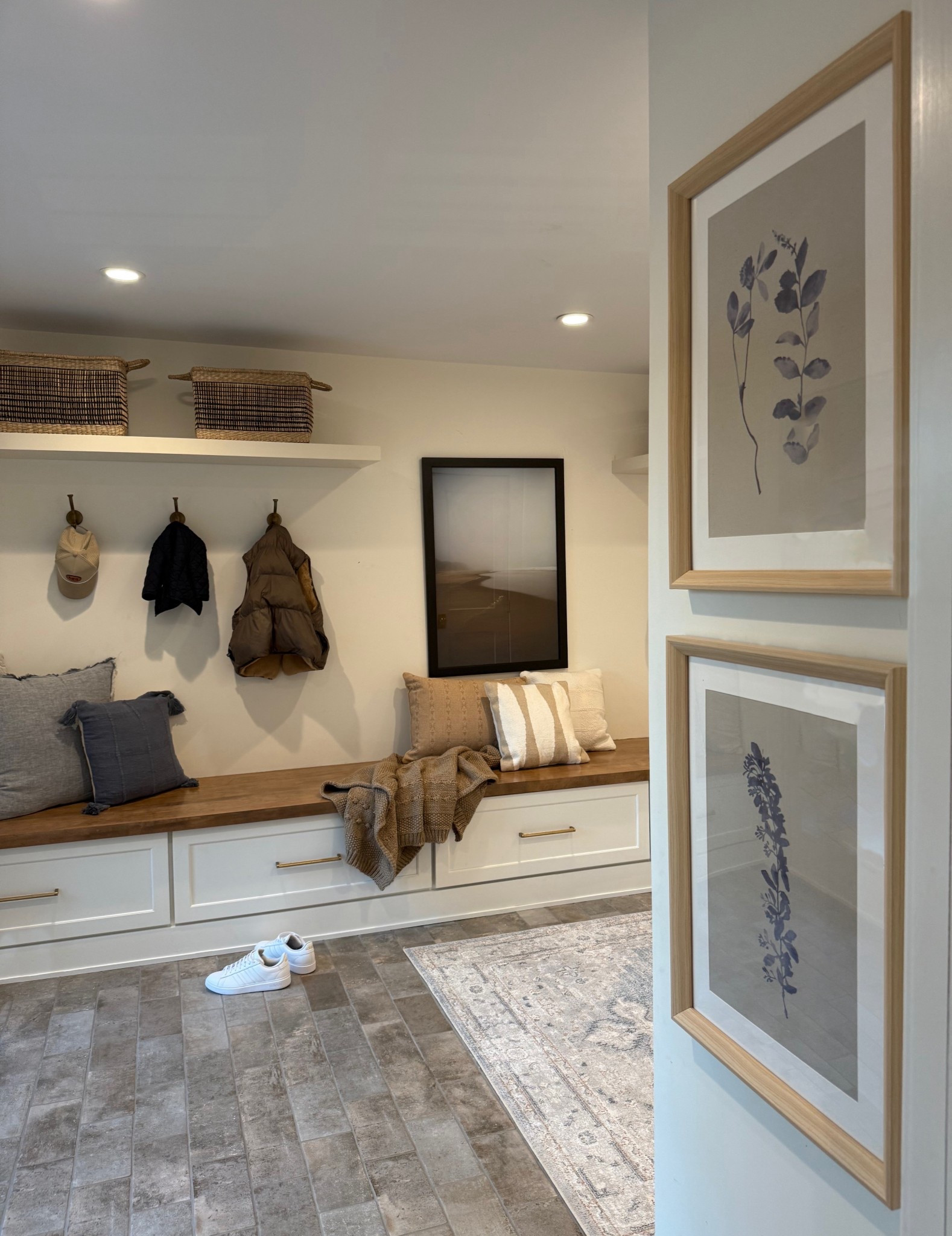 A view of the mudroom from the kitchen entrance highlights its seamless blend of style and utility. The rich blue stone floors flow effortlessly into the space, where a long bench with built-in storage anchors the room. Above, open shelves and neatly arranged hooks offer practical organization with a polished look. The warm tones, thoughtful design, and a striking photo on display make this practical area feel just as curated and inviting as the rest of the home.
