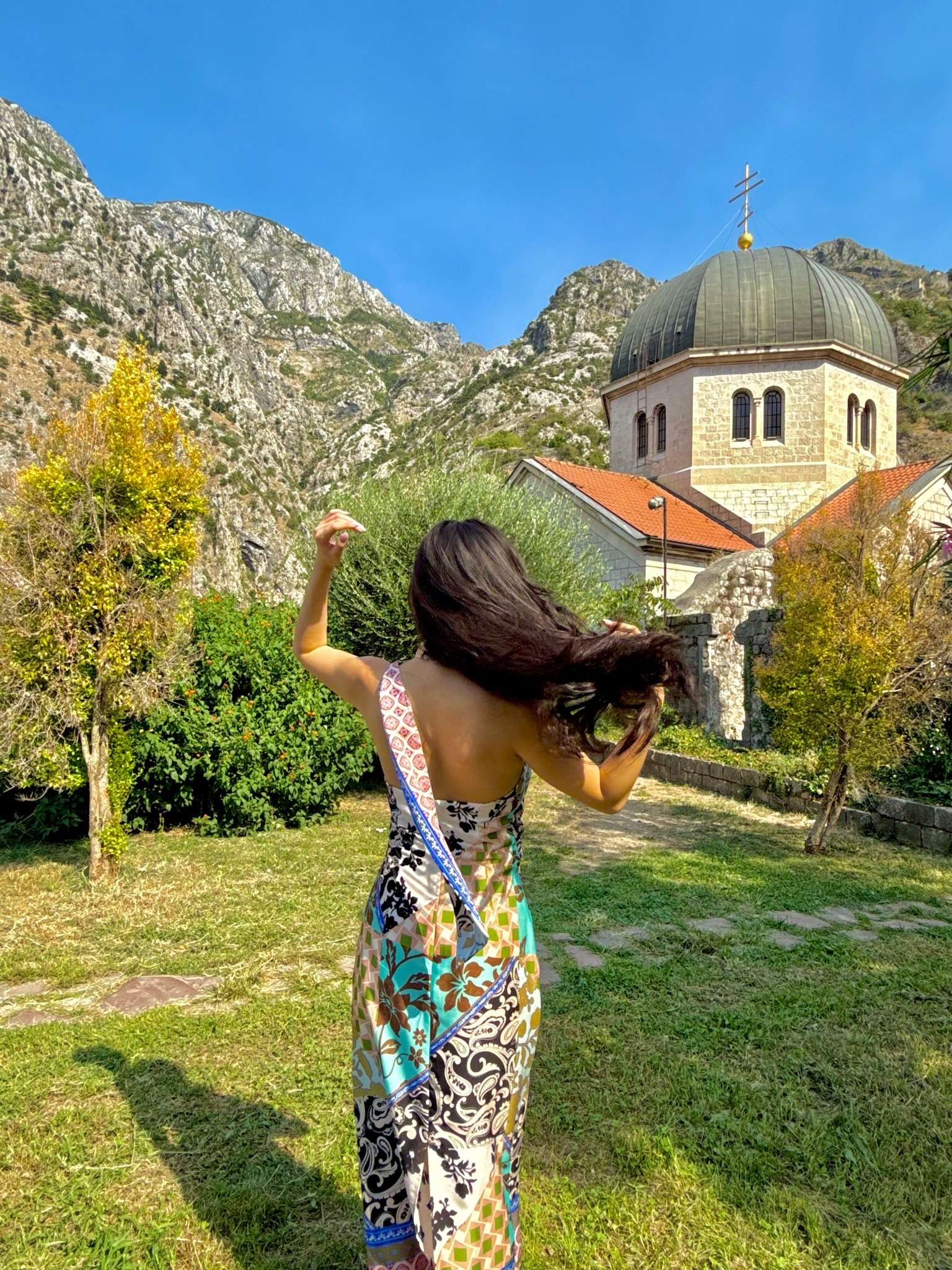 a colorful patchwork print backless maxi dress with mixed paisley, floral, and geometric panels in turquoise, pink, black, and cream, styled for a sunny afternoon exploring the historic stone churches of kotor old town in montenegro. the bold patchwork print and breezy backless silhouette make this the perfect statement vacation dress. perfect for kotor day trips, montenegro vacations, balkan coast travels, and mediterranean summer getaways. shop this look linked above.