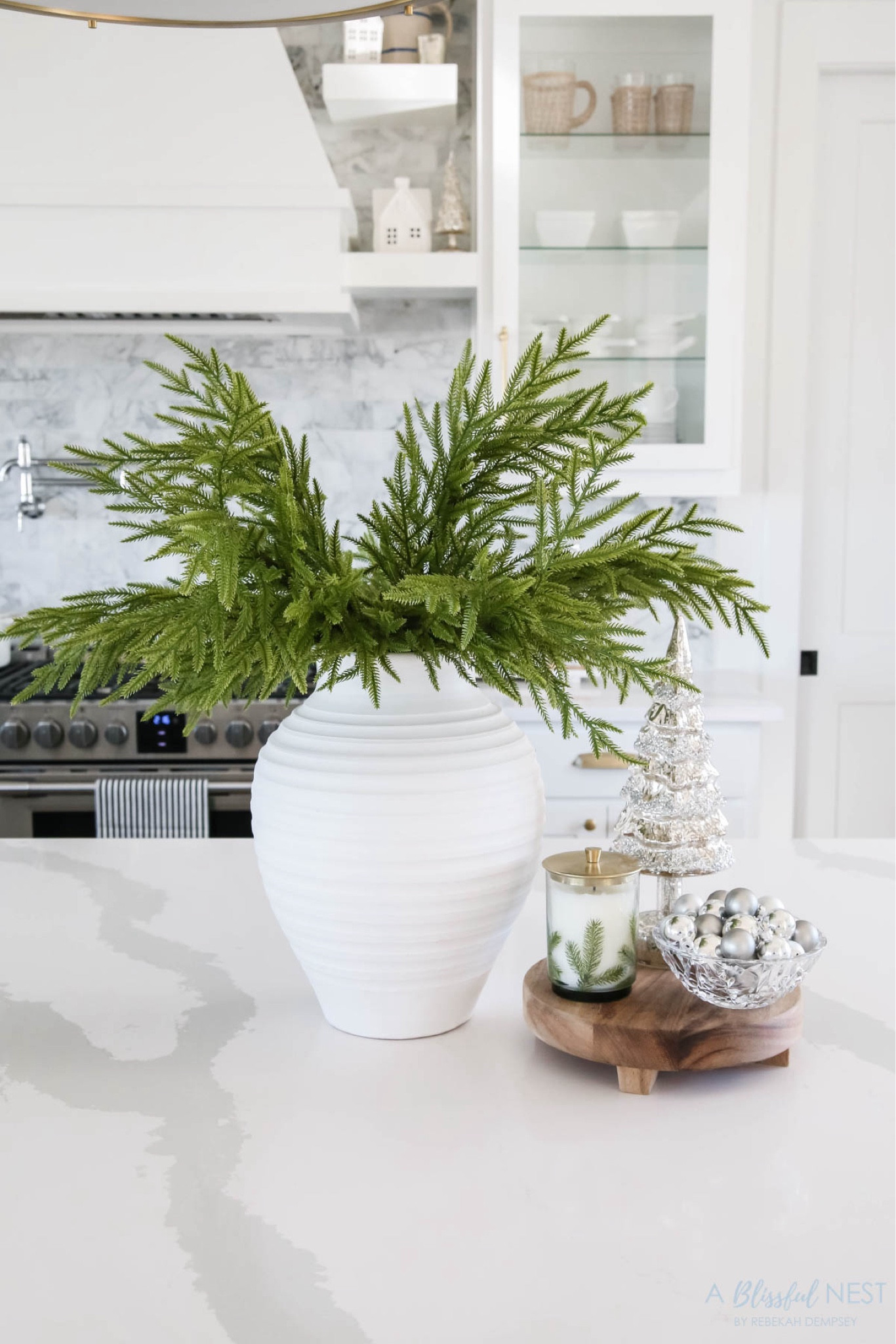 Holiday kitchen island details. Includes the vital Norfolk pine stems, ribbed white vase, wood pedestal, Fraser fir candle, and a bowl or ornaments. 

Holiday home tour, Christmas home tour, Christmas kitchen, white kitchen, coastal style, coastal grandmother

#LTKHome #LTKHoliday #LTKSeasonal
