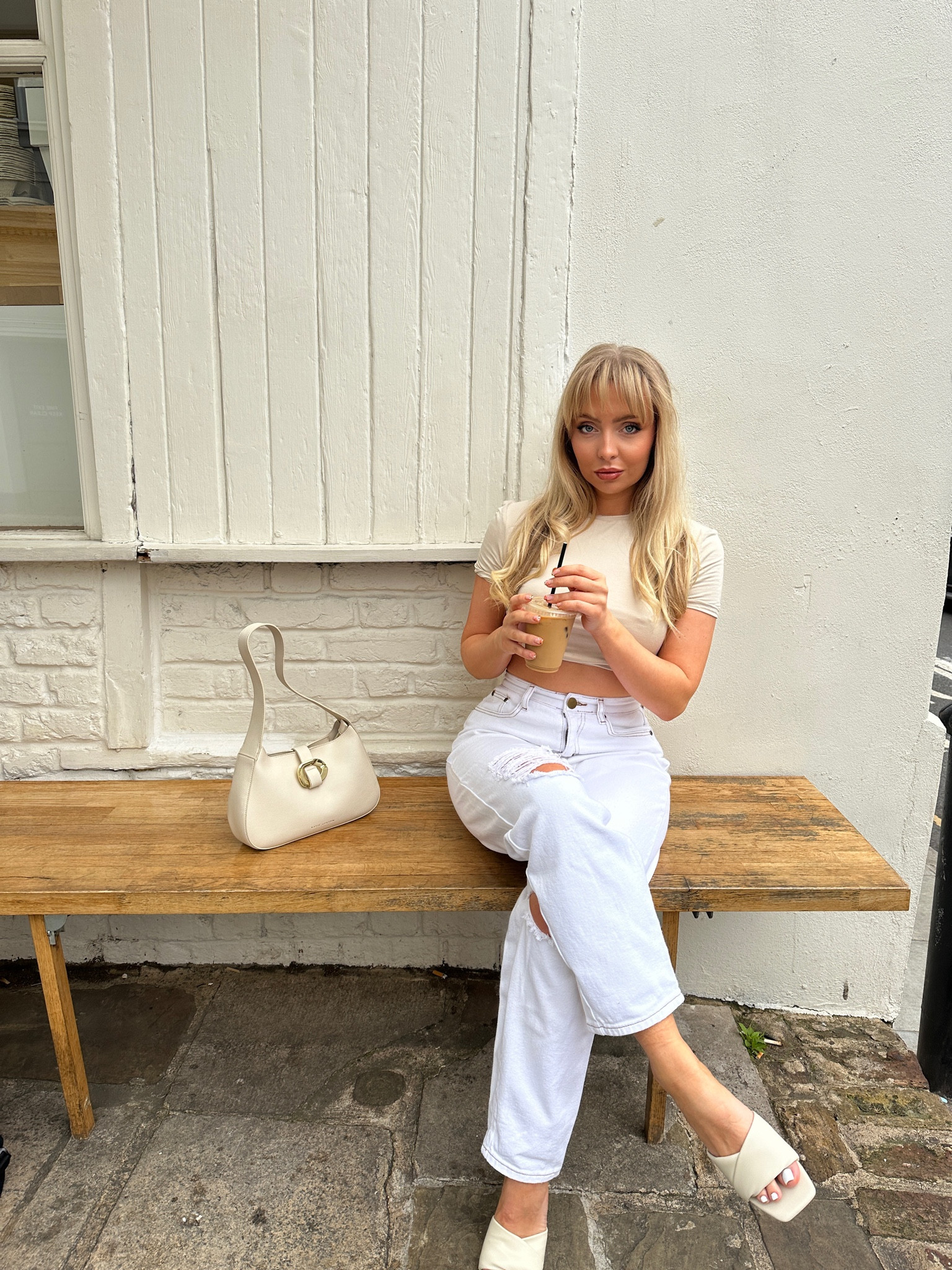Girl sat with a coffee wearing a cream cropped TShirt, white ripped jeans, and a cream Katie Loxton bag. She is wearing cream flat sandals  

#LTKunder50 #LTKFind #LTKunder100
