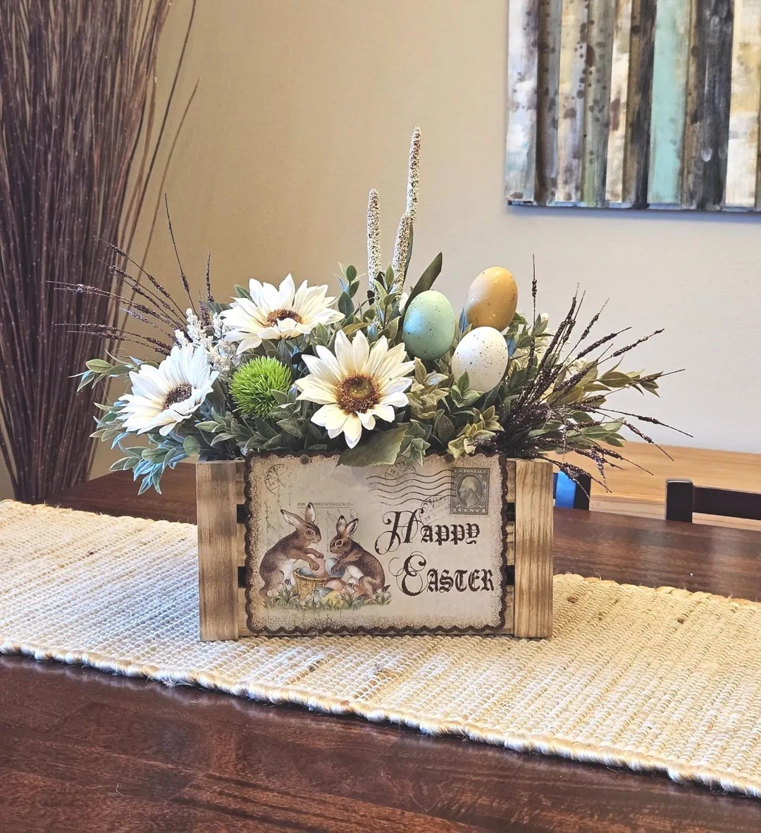 Celebrate Easter with this beautiful rustic centerpiece featuring soft neutral tones and natural textures that blend effortlessly with farmhouse décor. Designed in a wooden crate, the arrangement showcases cream sunflowers, speckled Easter eggs in brown, seafoam green, and white, along with berry accents, tall textured stems, and lush greenery. A sparkling brown accent spray and a charming “Happy Easter” postcard on the crate complete the design, creating a one-of-a-kind display that looks stunning on a table, entryway, or mantel. #easterdecor #springdecor #farmhousestyle #rusticdecor #centerpieceideas #ltkhome #homedecorfinds

#LTKSeasonal #LTKHome