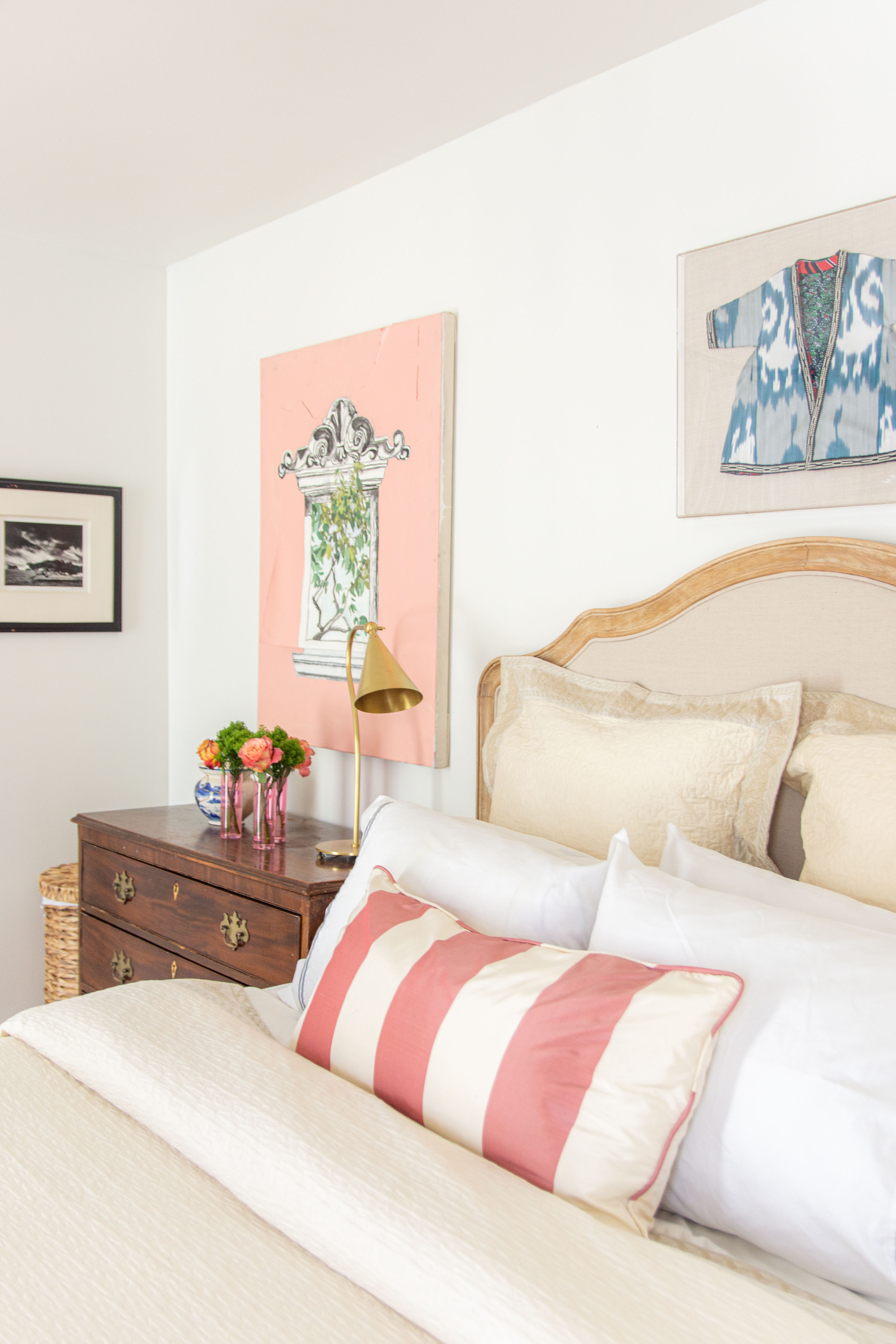 Bright and cozy bedroom inspiration featuring neutral bedding layered with crisp white pillows, a striped lumbar pillow in blush pink and cream, and a soft quilted duvet. A classic wooden headboard pairs beautifully with vintage-inspired nightstands styled with fresh flowers, a gold reading lamp, and framed wall art for a collected look. Perfect bedroom decor ideas for creating a timeless, elegant space with a touch of modern charm. 

 