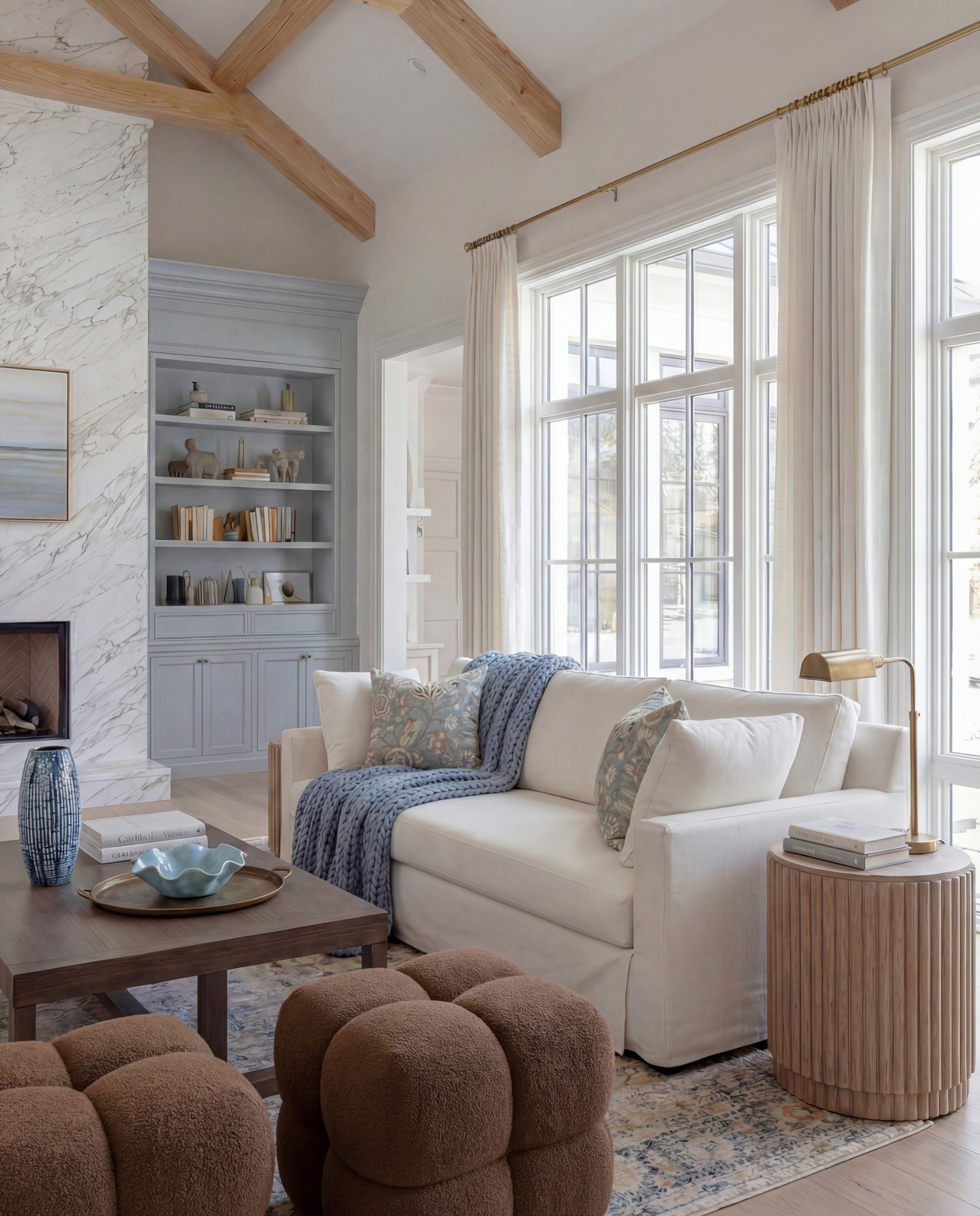 Shop this look:
• Exposed wood ceiling beams
• Blue floral wallpaper (ceiling + vaults)
• Marble fireplace surround
• Gas fireplace insert
• Framed neutral landscape artwork
• Built-in bookcases with lower cabinetry (soft blue-gray)
• Styled books + neutral ceramic decor
• Cream upholstered sofa
• Blue chunky knit throw blanket
• Blue and neutral accent pillows (patterned + solid)
• Upholstered accent chairs with wood frames
• Brown leather ottomans
• Rectangular wood coffee table
• Blue ceramic decorative vase
• Blue decorative bowl
• Stacked coffee-table books
• Light blue patterned area rug
• Small round wood side table
• Table lamp with white shade
• Floor-to-ceiling windows with sheer white curtains

#LTKHome #LTKFindsUnder100 #LTKSaleAlert