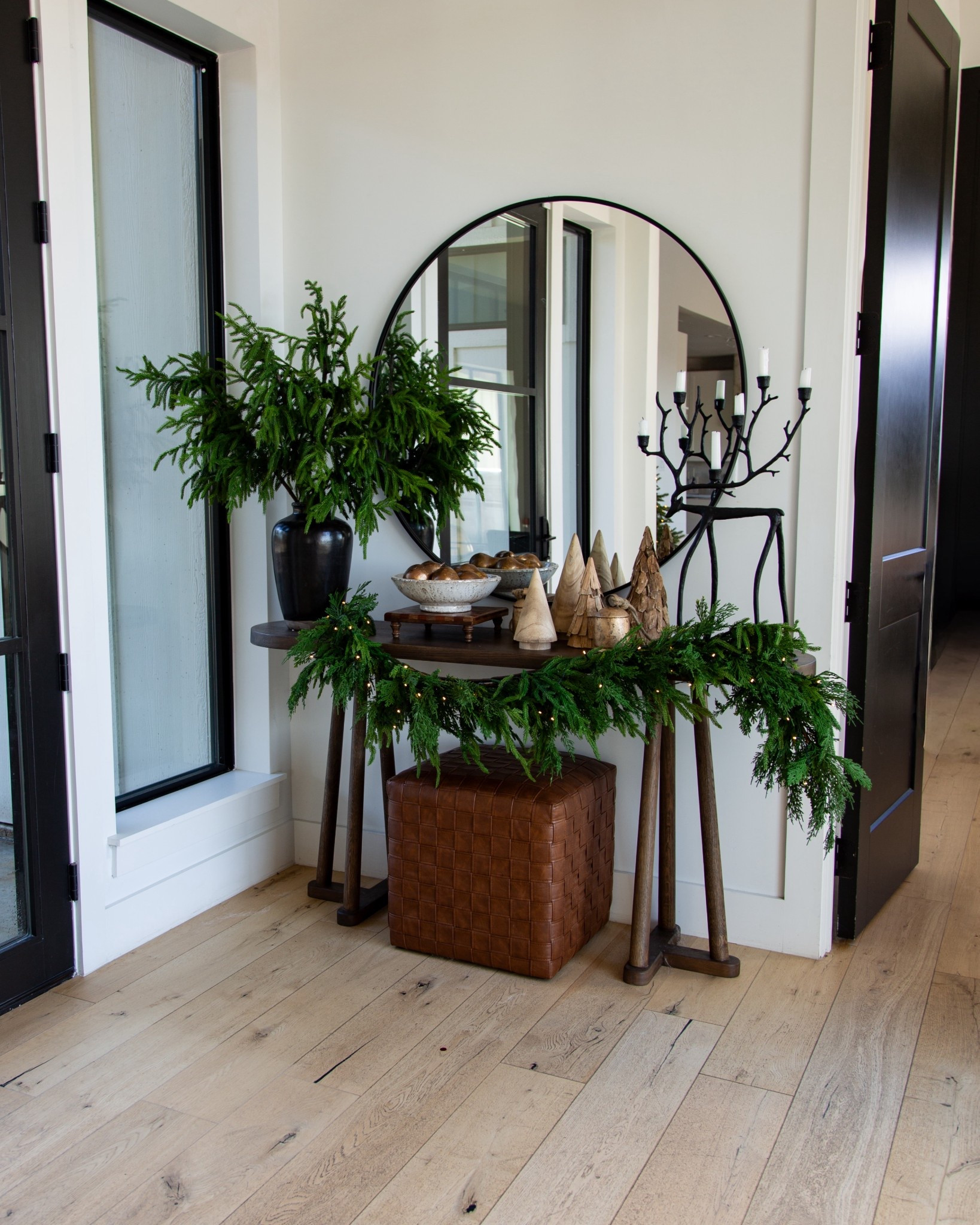 A little holiday moment in our entryway. I kept it simple with this oval black mirror, wood console table, and my favorite woven leather ottoman tucked underneath for extra texture. The garland with warm lights, wood Christmas trees, and black candelabra add just enough holiday charm without feeling overdone. I also brought in a big greenery arrangement in a black ceramic vase to keep everything looking fresh and full.


#LTKHoliday #LTKHome #LTKGiftGuide