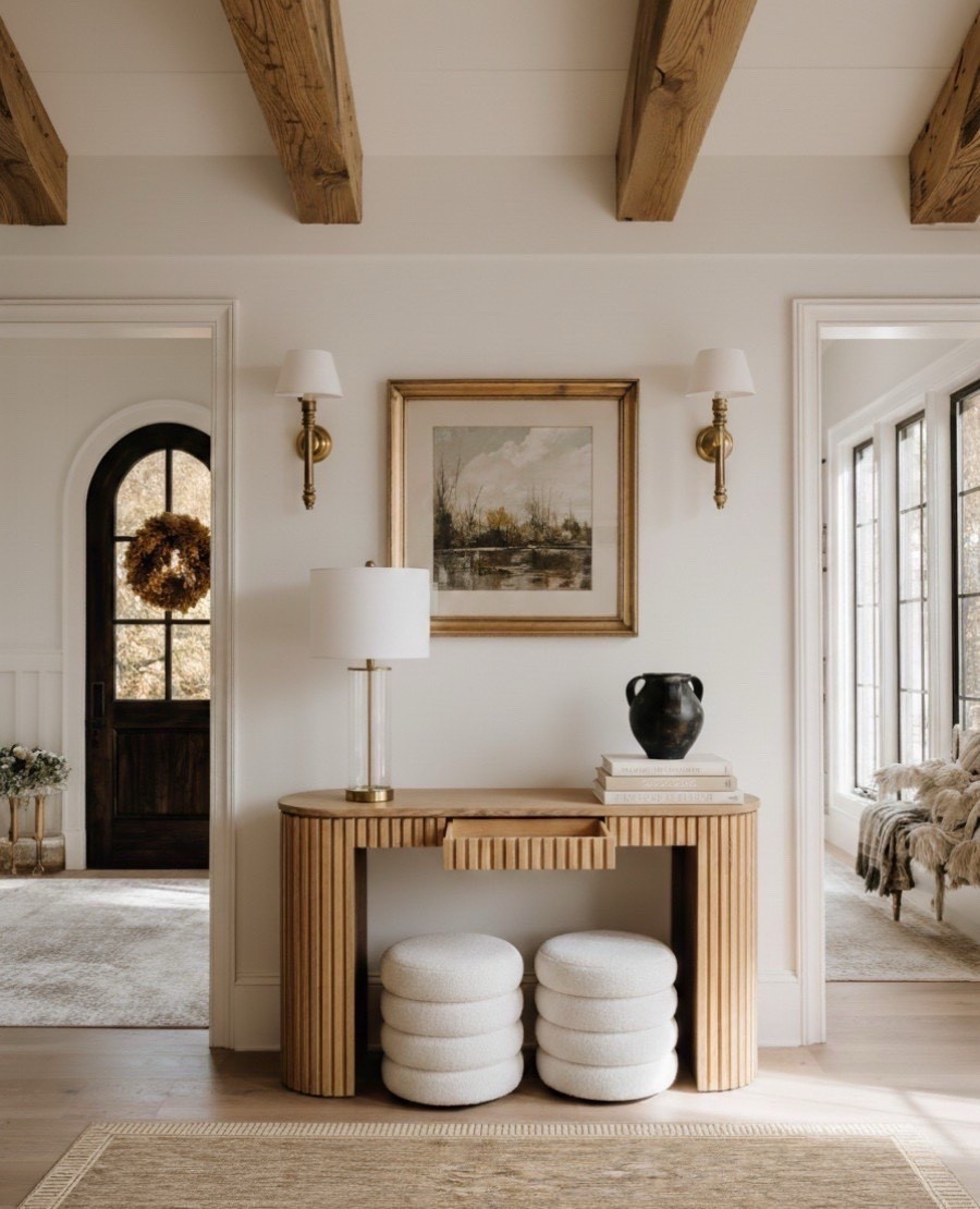 This beautifully styled neutral foyer blends organic textures and timeless details for a warm, inviting entryway. The fluted wood console table anchors the space with natural warmth, while the scenic landscape wall art in a gold frame adds a classic touch. Paired with a glass tube brass table lamp, modern gold wall sconces, and bouclé round ottoman stools, every element brings softness and sophistication. Layered with a neutral patterned area rug and natural oak ceiling beams, this foyer creates the perfect first impression for your home.

#LTKSaleAlert #LTKHome