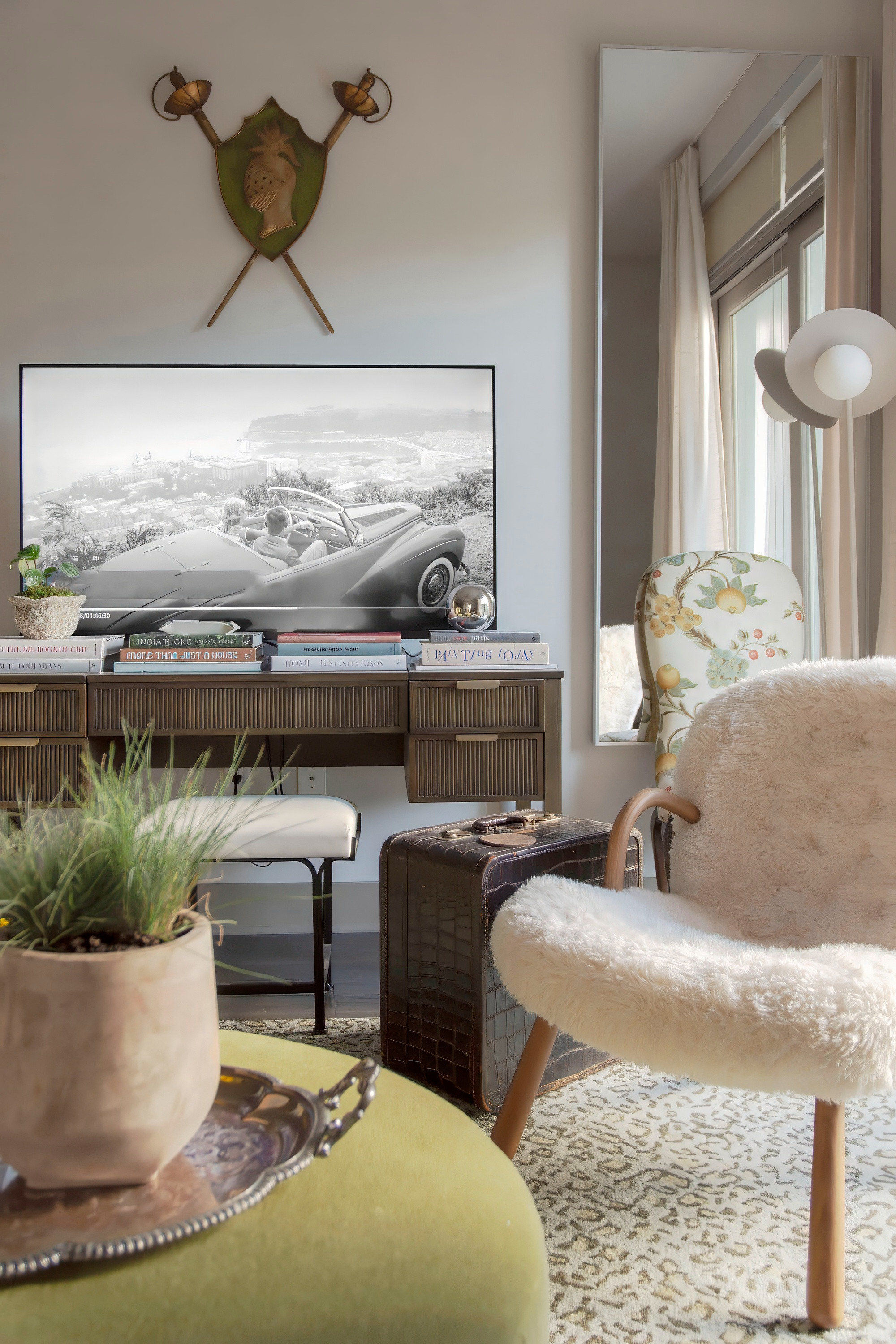 Neutral-toned living room with a cozy modern eclectic vibe. This stylish space features a boucle accent chair, a vintage floral armchair, and a textured green ottoman topped with a decorative tray and potted greenery. A sleek console table is styled with stacked coffee table books and small decor, while a wall-mounted TV blends into the design as framed art. Warm textures, layered textiles, and natural light complete this inviting home decor look, perfect for chic apartment living and timeless interior inspiration. 

 