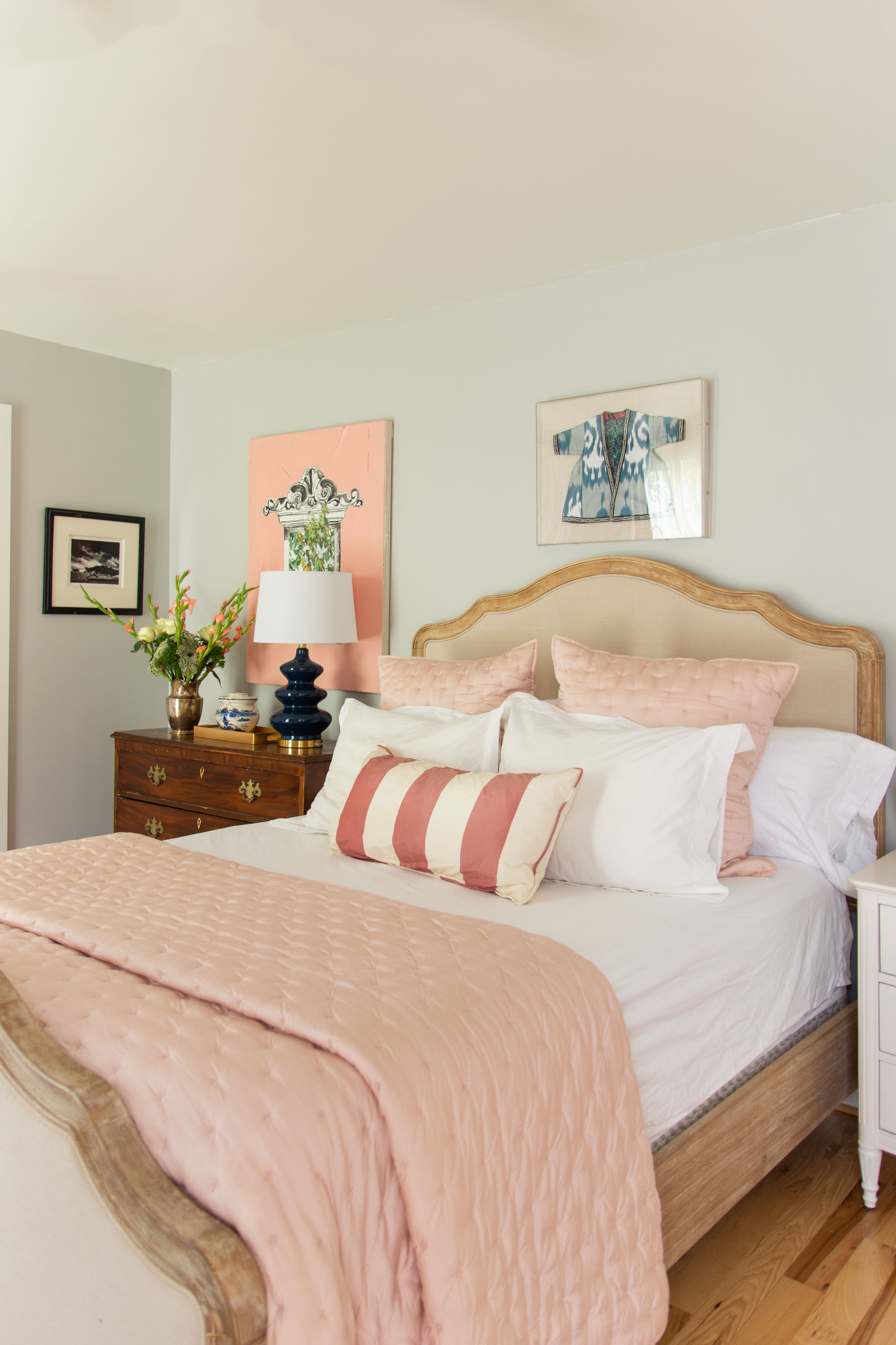 Soft and serene bedroom design featuring a neutral upholstered bed frame with natural wood trim, layered with blush pink bedding, tufted pillows, and a striped lumbar pillow for a pop of pattern. The light gray walls provide a calm backdrop, accented by eclectic wall art and a mix of classic and modern decor. A vintage wooden dresser topped with a navy ceramic lamp, floral arrangement, and decorative accessories adds warmth and character. Perfect example of blending traditional furniture with fresh, feminine touches for a cozy, stylish retreat. 

 