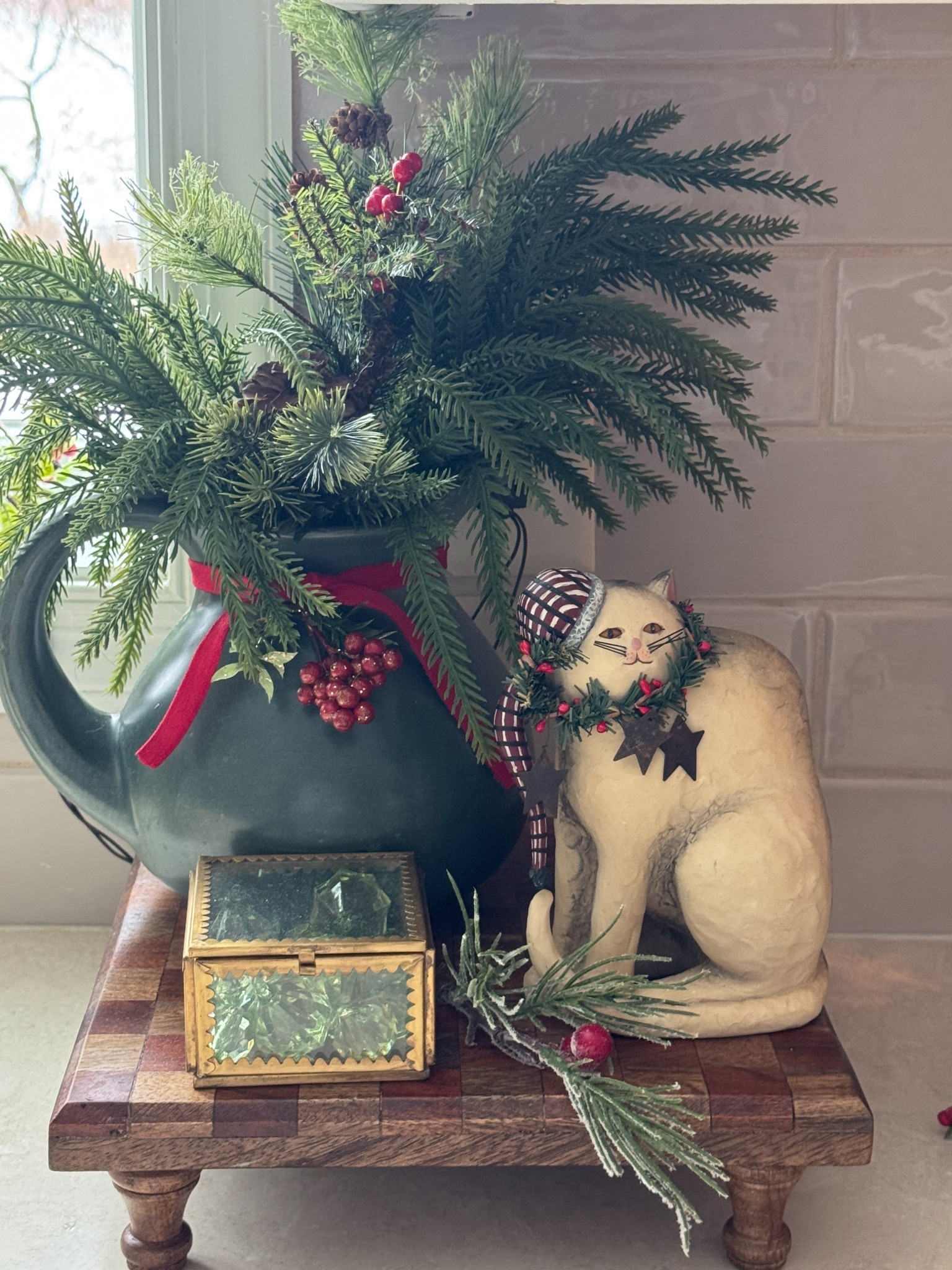 Christmas tray display: Checkered wood riser, matte stoneware pitcher, brass trinket box, similar cat figurine, greens, red ribbon, berry picks. 