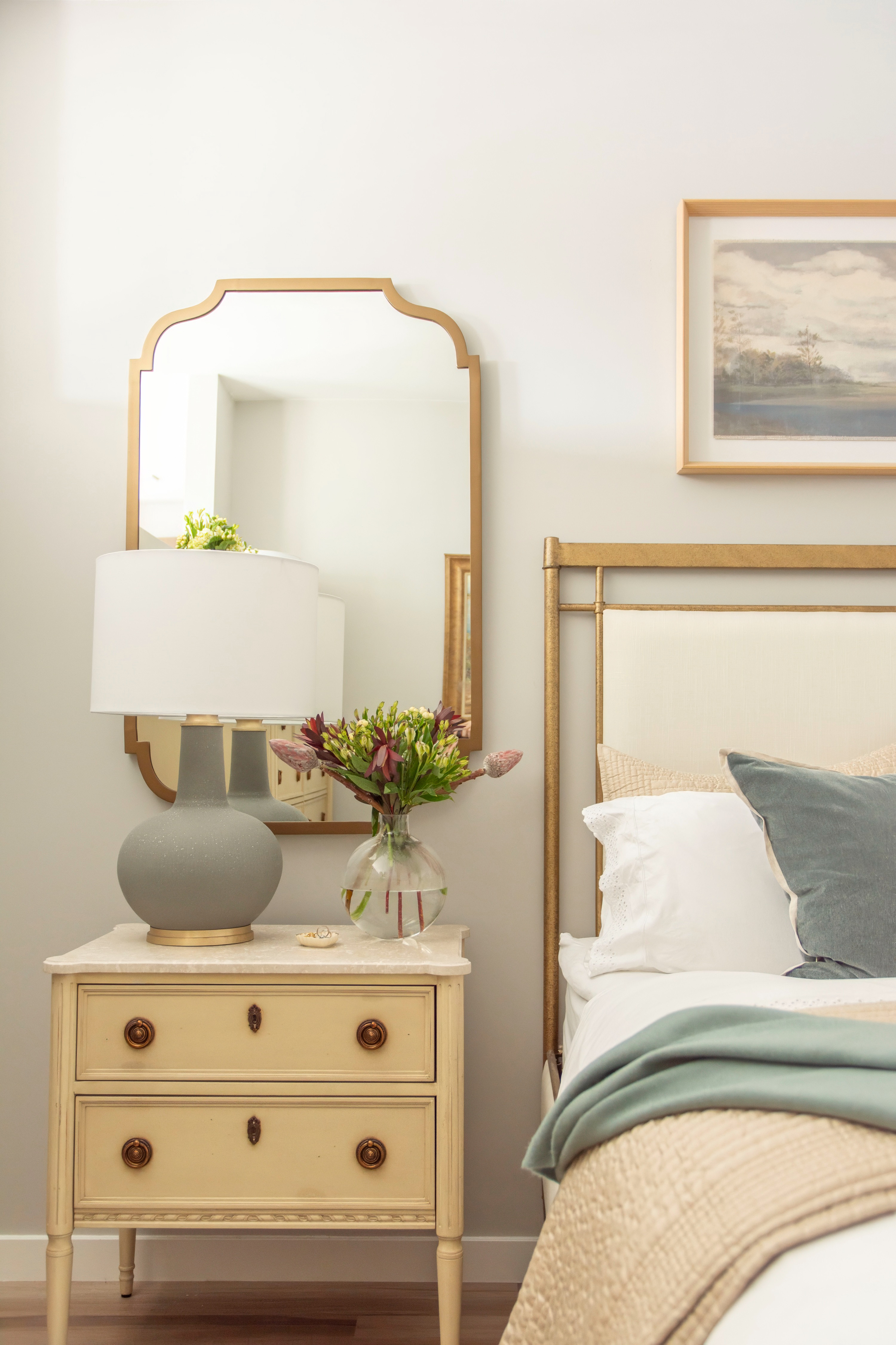 Cozy bedroom styling inspiration featuring a neutral nightstand with vintage-inspired brass knobs, paired with a classic ceramic table lamp topped with a crisp white shade. The layered bedding mixes soft white sheets, a muted sage green throw pillow, and a textured beige quilt for a timeless look. A gold framed mirror adds warmth and elegance above the nightstand, reflecting natural light and enhancing the space. Fresh florals in a simple glass vase bring a pop of greenery and seasonal charm. Perfect blend of modern farmhouse and traditional bedroom décor. 

 