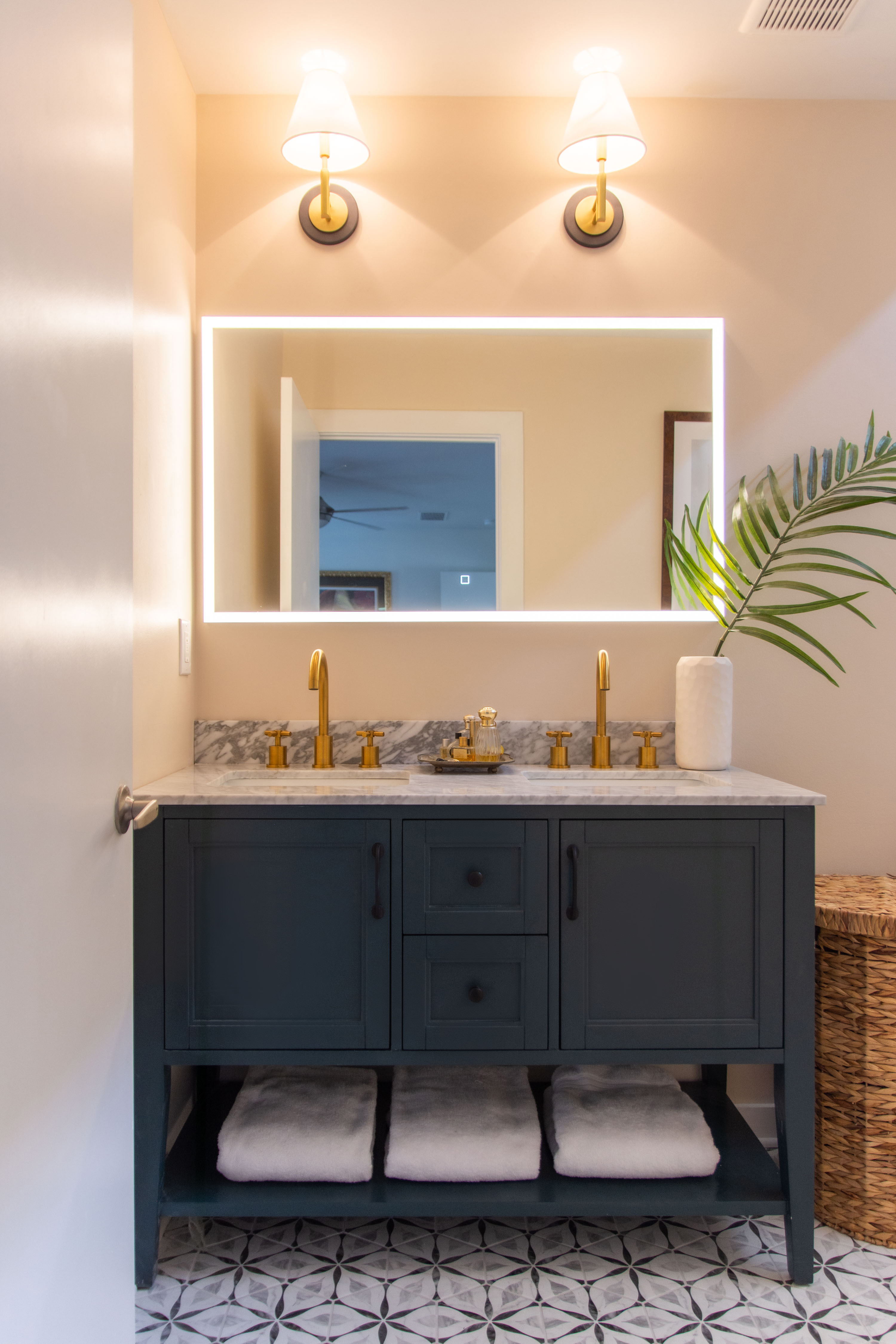 Modern primary bathroom design featuring a double vanity with marble countertop, brass fixtures, and a navy blue cabinet. The backlit LED mirror and gold wall sconces create a warm and inviting glow, while the geometric patterned tile adds a stylish touch. Open shelving with neatly folded white towels and a woven hamper keep this space both functional and elegant. Perfect inspiration for anyone looking for bathroom remodel ideas, vanity styling tips, or ways to mix brass hardware with marble countertops. 

 