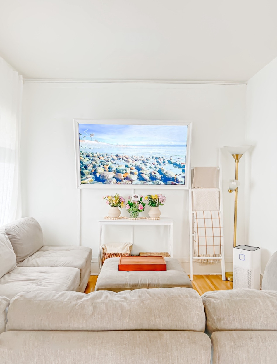 Warm and minimal living room with neutral colors gray couch, beach scene on TV white ladder with cozy, blankets, white entryway table under the TV, air purifier, lighting, clean

#LTKunder100 #LTKFind #LTKhome