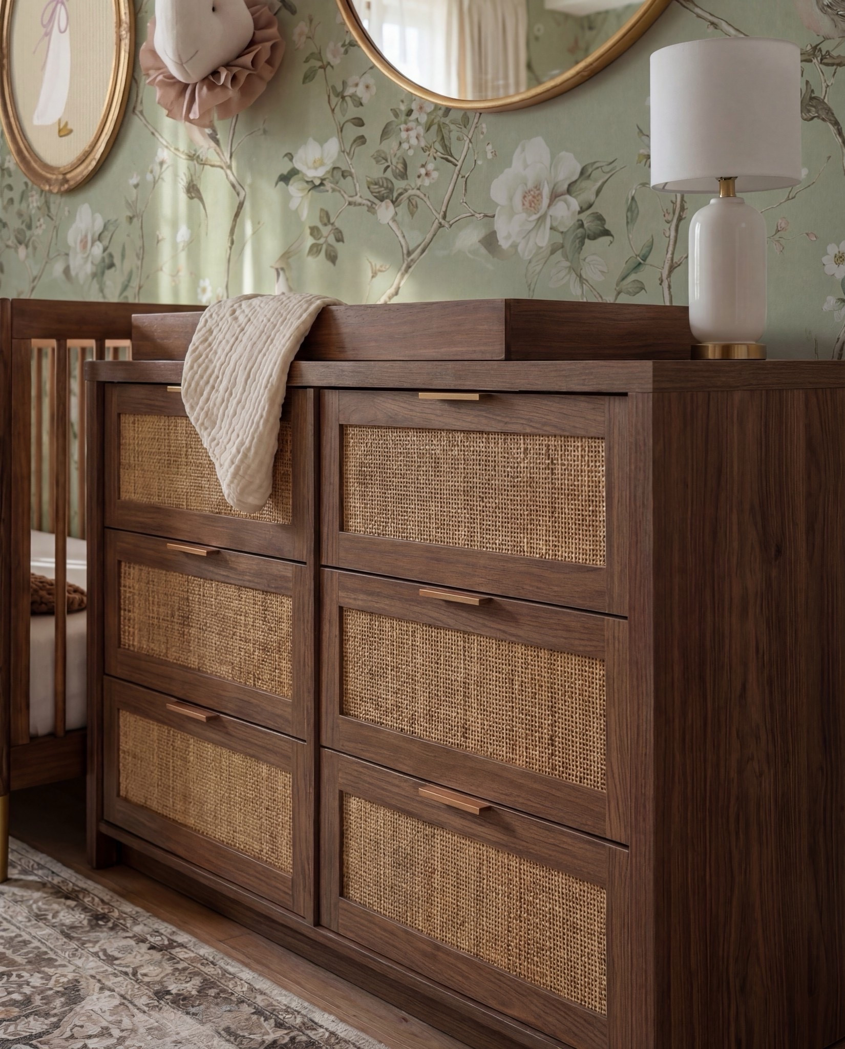 Shop the Look:
• Floral wallpaper (soft green with botanical print)
• Solid wood crib (warm walnut tone)
• Knit throw blanket (neutral brown)
• Wood dresser with woven drawer fronts
• Changing tray (wood)
• Round brass-framed wall mirror
• Oval framed animal artwork
• Decorative wall animal mount
• Upholstered accent chair (ivory)
• Green accent pillows
• Floor lamp (brass base, white shade)
• Table lamp (ceramic base, white shade)
• Pendant ceiling light (soft white globe)
• Neutral vintage-style area rug
• Light wood flooring
• Sheer curtains with linen drapery panels

#LTKHome #LTKSaleAlert