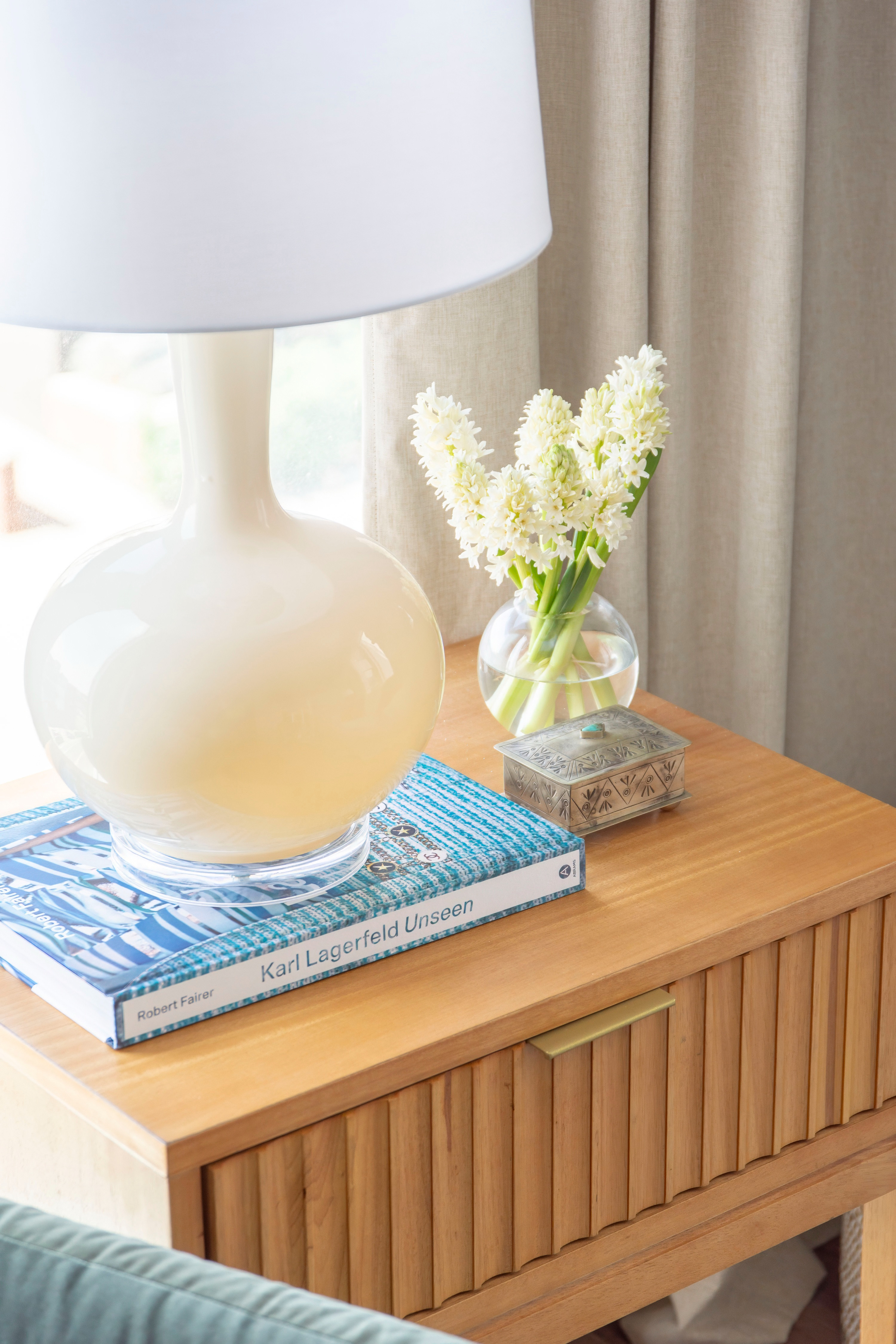 A chic and modern nightstand styled with timeless details. This wood bedside table features a ribbed drawer design with a sleek brass pull, perfectly complemented by a glossy white ceramic lamp with a simple round base. For a touch of freshness, a clear glass vase holds white hyacinth flowers, while a decorative silver trinket box adds a vintage accent. Stacked beneath the lamp is a coffee table book, Karl Lagerfeld Unseen by Robert Fairer, bringing a fashion-forward element to the bedside look. This nightstand styling idea blends neutral tones, fresh florals, and designer coffee table books for a sophisticated bedroom setup. 

 