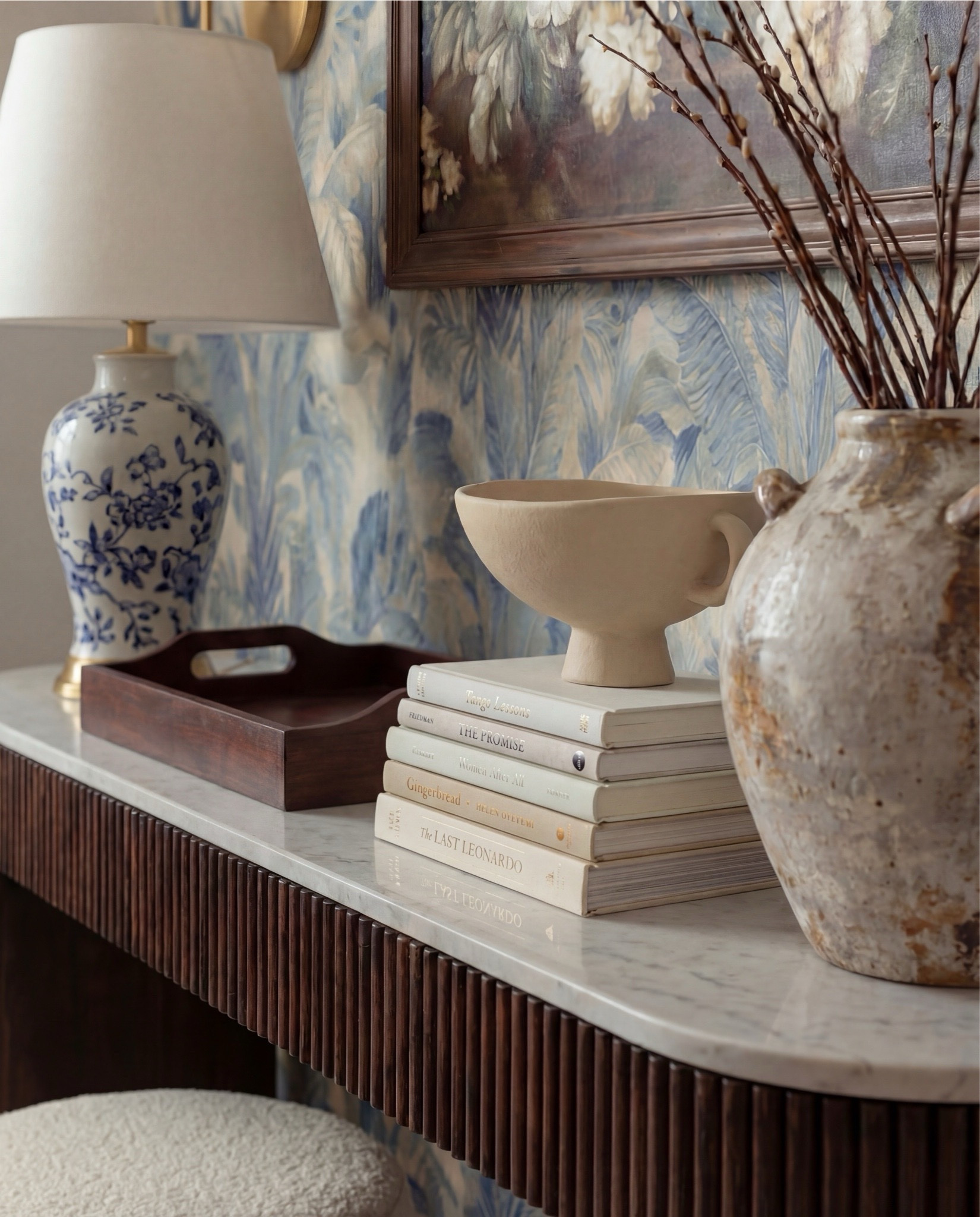 Shop the Look:
• Blue botanical wallpaper (leaf motif, muted navy + beige)
• Solid wood console table (dark walnut finish)
• Fluted wood console table legs
• White marble console tabletop
• Framed vintage floral wall art (dark wood frame)
• Brass wall sconces with white globe shades
• Blue and white ceramic table lamp
• White fabric lamp shade (tapered)
• Wooden decorative tray (rectangular)
• Stacked neutral coffee table books
• Sculptural ceramic bowl (cream stoneware)
• Distressed ceramic vase (antique white)
• Dried branches or twigs arrangement
• Bouclé ottomans (ivory, round)
• Light wood flooring
• White baseboards and trim

#LTKSaleAlert #LTKHome