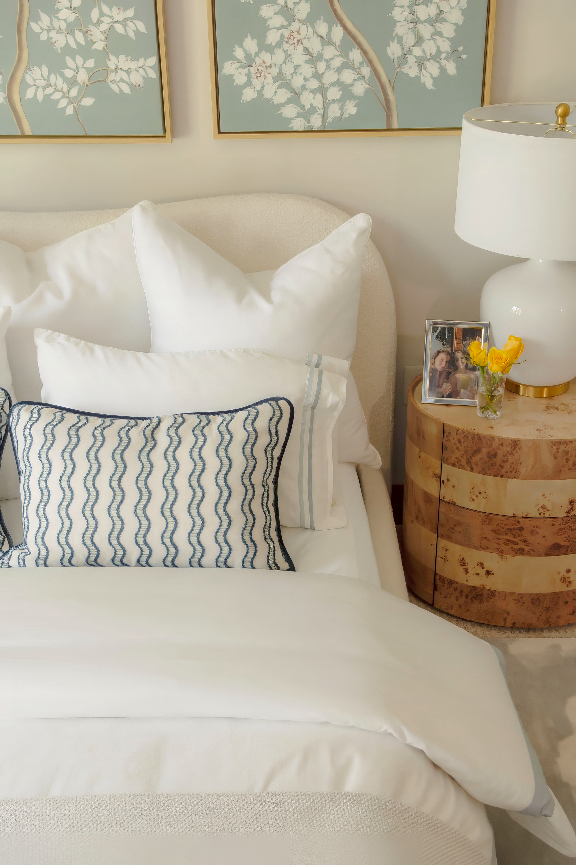 A cozy yet sophisticated bedroom refresh featuring layered white bedding with crisp euro shams and a textured accent pillow in a navy wave pattern. The look is paired with a modern burl wood nightstand, a chic white ceramic lamp with brass details, and a pop of color from fresh yellow tulips. Above the bed, botanical artwork in muted tones adds a soft, calming vibe. This design blends timeless neutrals with organic textures, creating an inviting space perfect for everyday comfort and relaxation. Shop similar bedding, pillows, nightstands, and decor to recreate this serene bedroom style. 

 