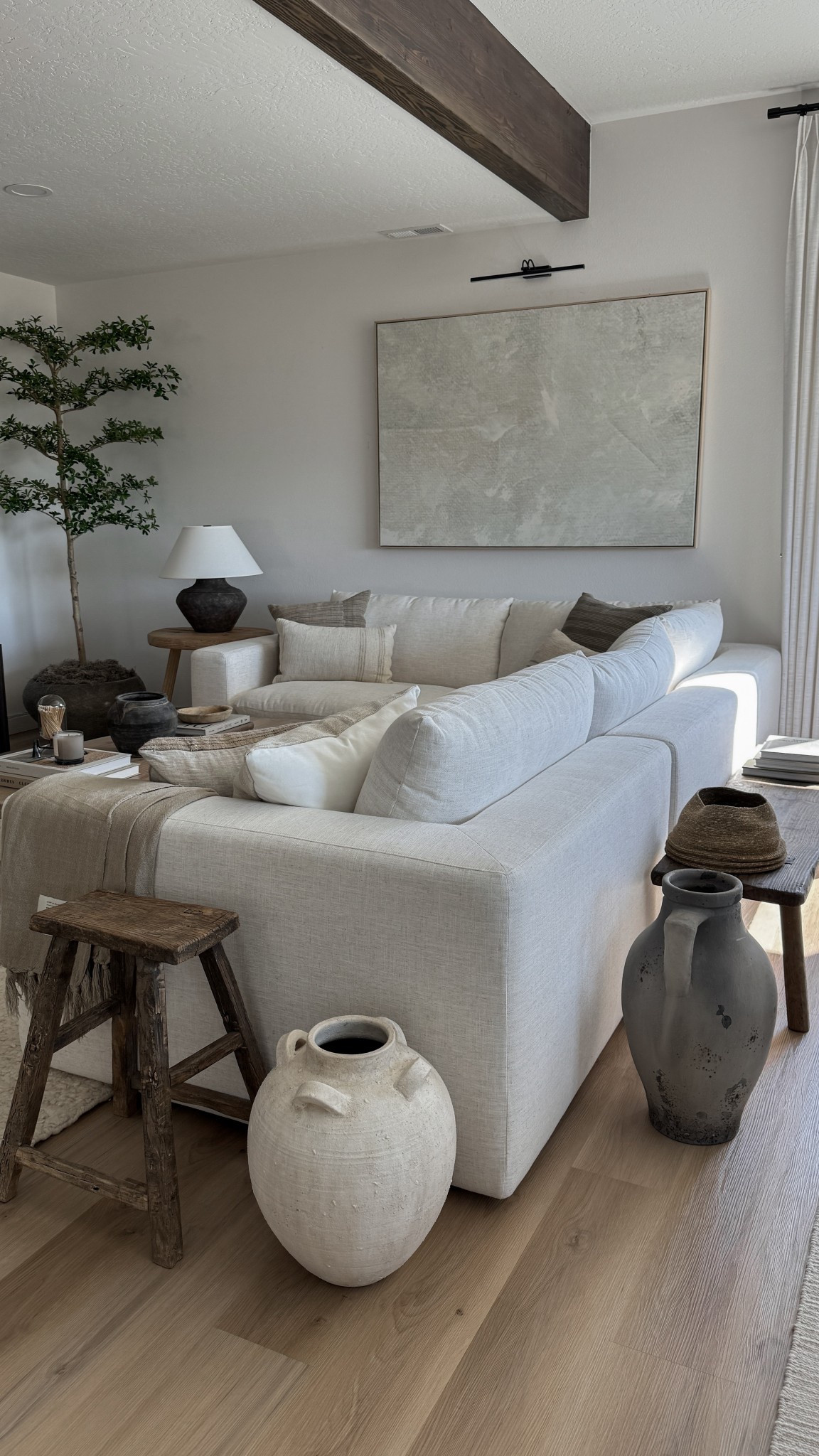 Neutral organic modern living room with cozy earthy decor details. This space features a large white sectional sofa styled with neutral throw blankets and textured pillows, rustic wood stools, and oversized ceramic vases for an earthy room aesthetic. The organic modern coffee table is styled with layered coffee table books and decor bowls, while the large wall art and faux olive tree bring in warmth and balance. Perfect inspiration if you’re looking for neutral home decor ideas, earthy living room inspiration, or organic modern home style. 

 #LTKHome