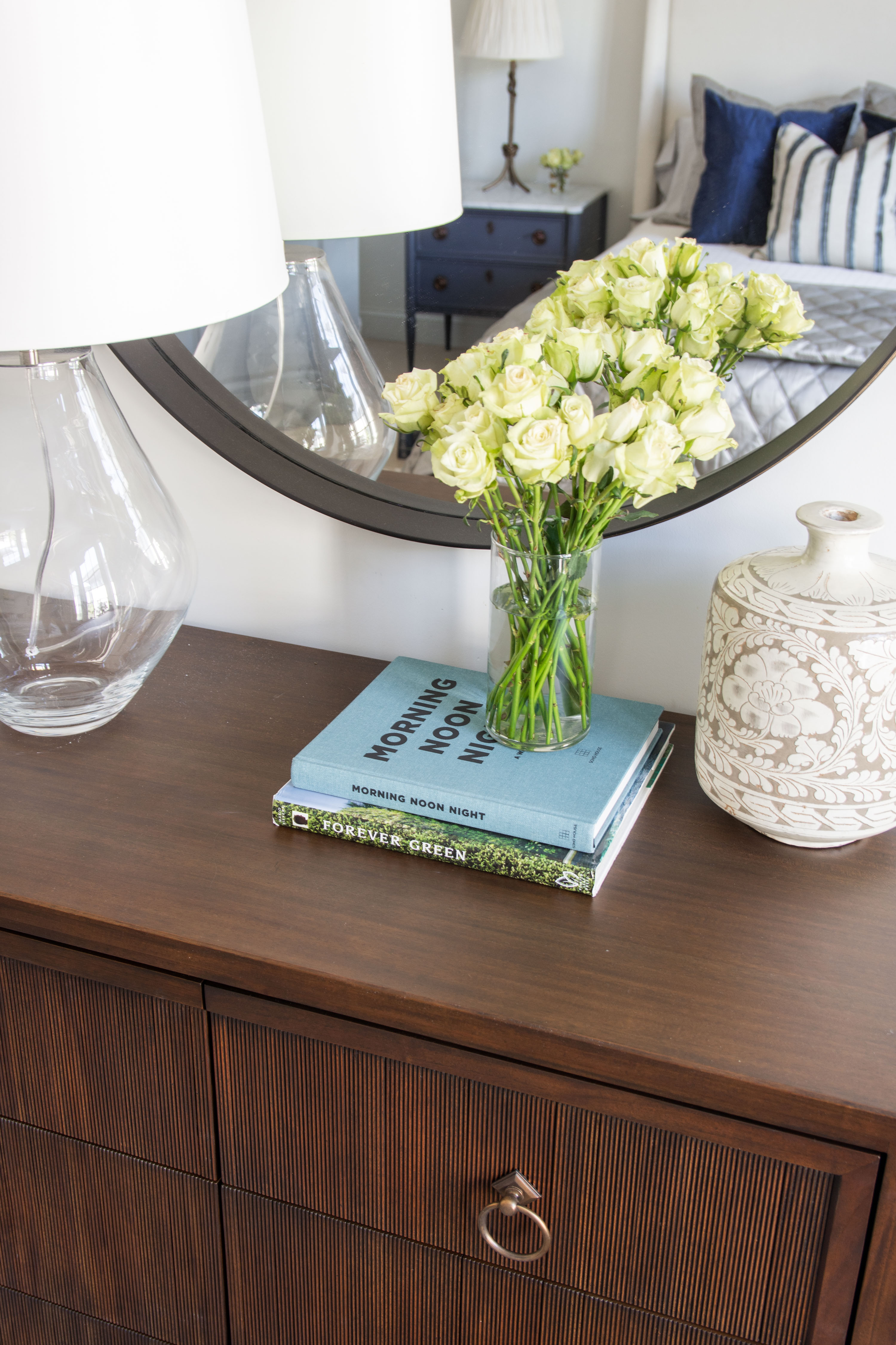 Chic bedroom styling with timeless details. This dark wood dresser is styled with a round mirror, glass lamp, and a white ceramic vase for a clean transitional look. A fresh bouquet of cream roses adds a soft, romantic touch, while stacked coffee table books — Morning Noon Night and Forever Green — bring in layered sophistication. Perfect modern farmhouse meets classic coastal vibes. Bedroom decor, dresser styling, home accessories, and interior design inspiration all in one polished setup. 

 