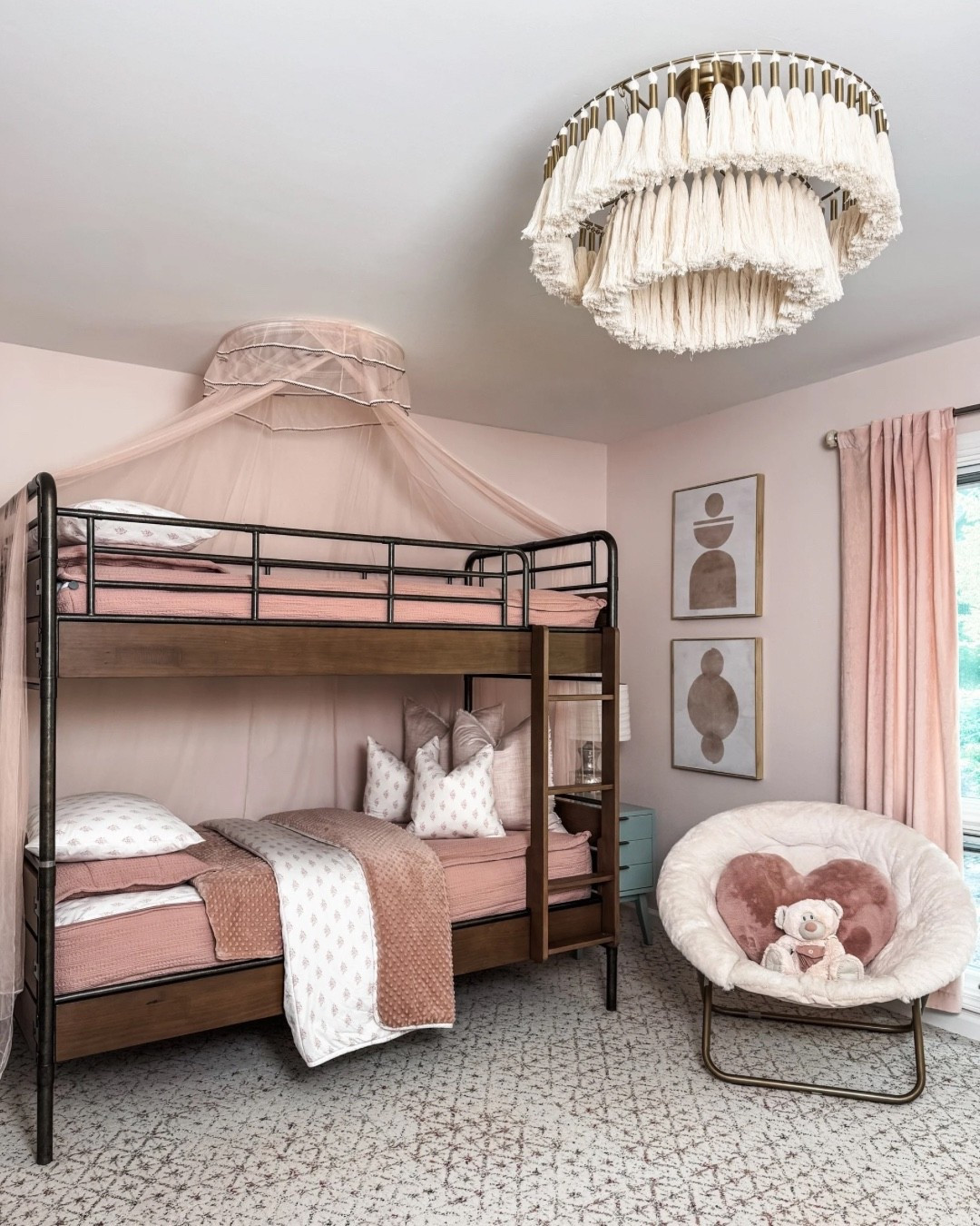 A dreamy kids’ bedroom ✨ This space features a wooden bunk bed layered with pink bedding and plush throw pillows, topped with a cozy canopy. The tassel chandelier adds texture overhead, while modern side tables with lamps and soft curtains frame the room. A cozy accent chair with heart pillow, framed wall art, and a patterned carpet by FLOR complete the look. 🌸

Girls: 
Alexandria Luxe Beddy’s (Twin, Minky, ZipBase)
Eden Blanket 
Ella Medium Pillow Cover 
Ella Pillowcase 
Ella Sham 
Eden Euro Pillow Cover 

#KidsRoomDesign #BunkBedStyle #PinkBedding #ThrowPillows #CanopyBed #TasselChandelier #SideTables #TableLamps #WallArt #AccentChair #PatternedRug #eanesinteriors

#LTKKids #LTKStyleTip #LTKFamily