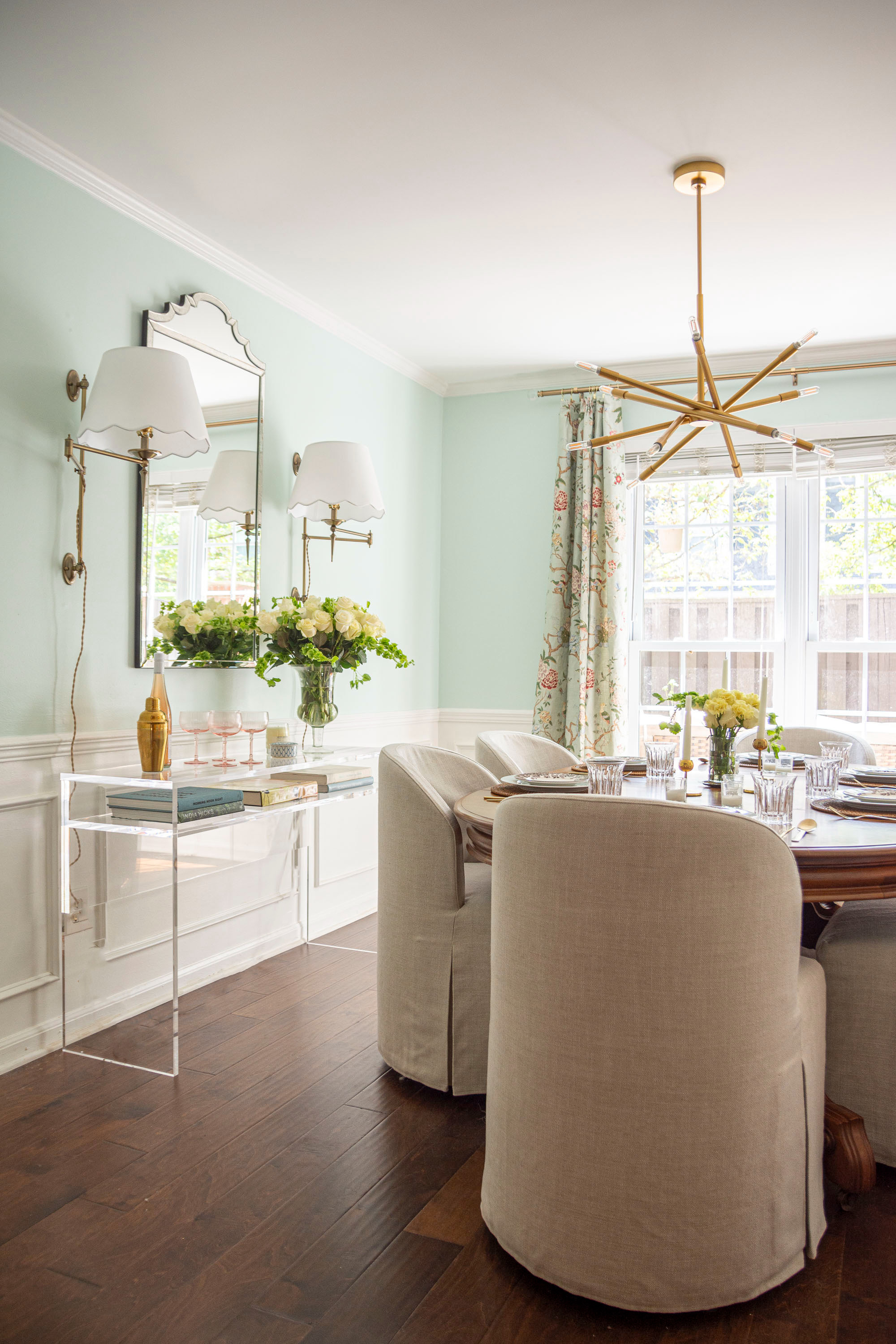 Bright and inviting dining room inspiration featuring soft mint green walls, classic white wainscoting, and a mix of modern and traditional details. A round wood dining table is styled with neutral upholstered chairs, crystal glassware, and fresh florals for an elegant tablescape. The gold starburst chandelier adds a bold mid-century touch, while the acrylic console table keeps the space airy and chic. Layered accents like a scalloped mirror, floral drapery panels, and brass swing-arm sconces complete the look. Perfect blend of timeless style and modern sophistication for a cozy yet polished dining space. 

 