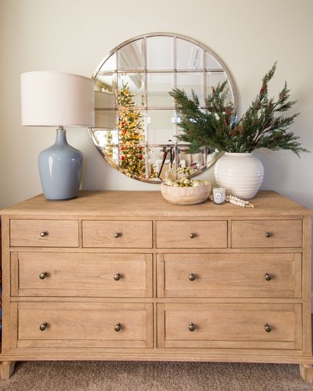Our light wood dresser with large white vase and faux greenery. Other items include wood bowl with gold ornaments, wood necklace, scented candle and round silver mirror with square beveled glass inside. Reflecting in the mirror is a faux Christmas tree, a nightstand with wicker style doors and striped linen curtains. 

Christmas décor, Christmas bedroom, wall mirror, Christmas greenery, Christmas tree, pottery barn furniture, pottery barn dresser, tree collar, bedroom curtains, master bedroom Christmas, guest bedroom Christmas #ltkholiday #ltkfamily  

#LTKSeasonal #LTKstyletip #LTKunder50 #LTKunder100 #LTKhome #LTKsalealert #LTKHoliday #LTKhome #LTKstyletip