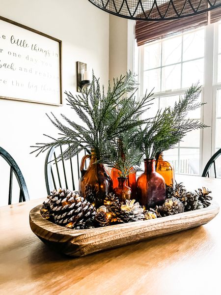 Make this easy winter centerpiece in less tha 15 minutes. Amber glass bottles filled with cypress and cedar greenery surrounded by pinecones in a wood dough bowl. Add some twinkle fairy lights for a cozy look all season long. #winterhomedecor #amberbottles #wintertablescape

#LTKunder50 #LTKhome #LTKSeasonal