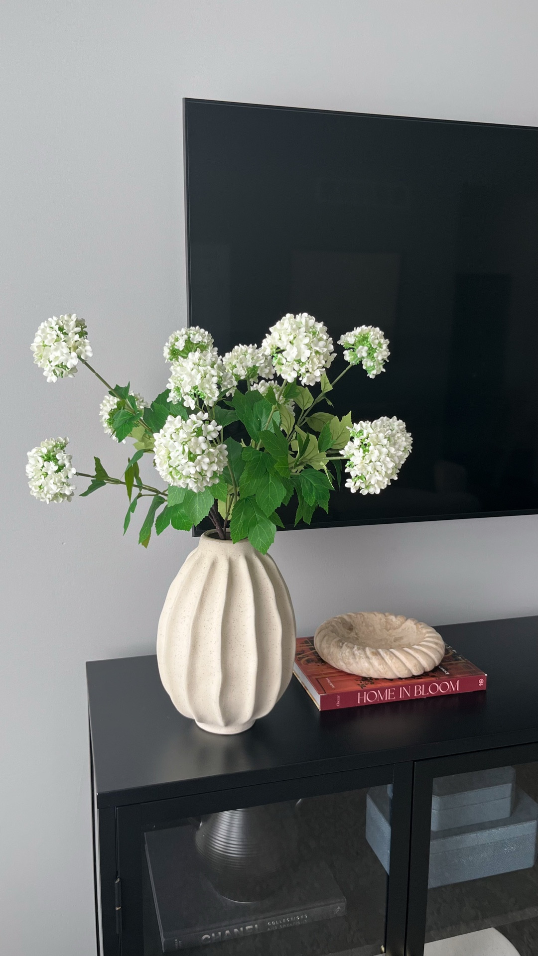 Console table spring refresh 🌸🍃✨ Faux silk hydrangea floral stems, wavy vase is old from Target (linking other similar styles!), Home in Bloom book, Studio McGee twisted edge cream marble dish, travertine stone book ends, other decorative home decor books, decorative boxes, candlesticks and candle stick holders, black and glass console table 

#LTKselfcare #LTKSeasonal #LTKHome