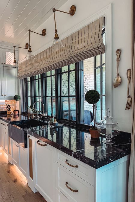 Kitchen sink styling with my fav essential oil hand soap, faux topiaries on sale, overhead bent arm sconces, polished nickel bridge faucet, and brass cabinet pulls. 
Roman shades from @uptowndrapes

#LTKsalealert #LTKhome