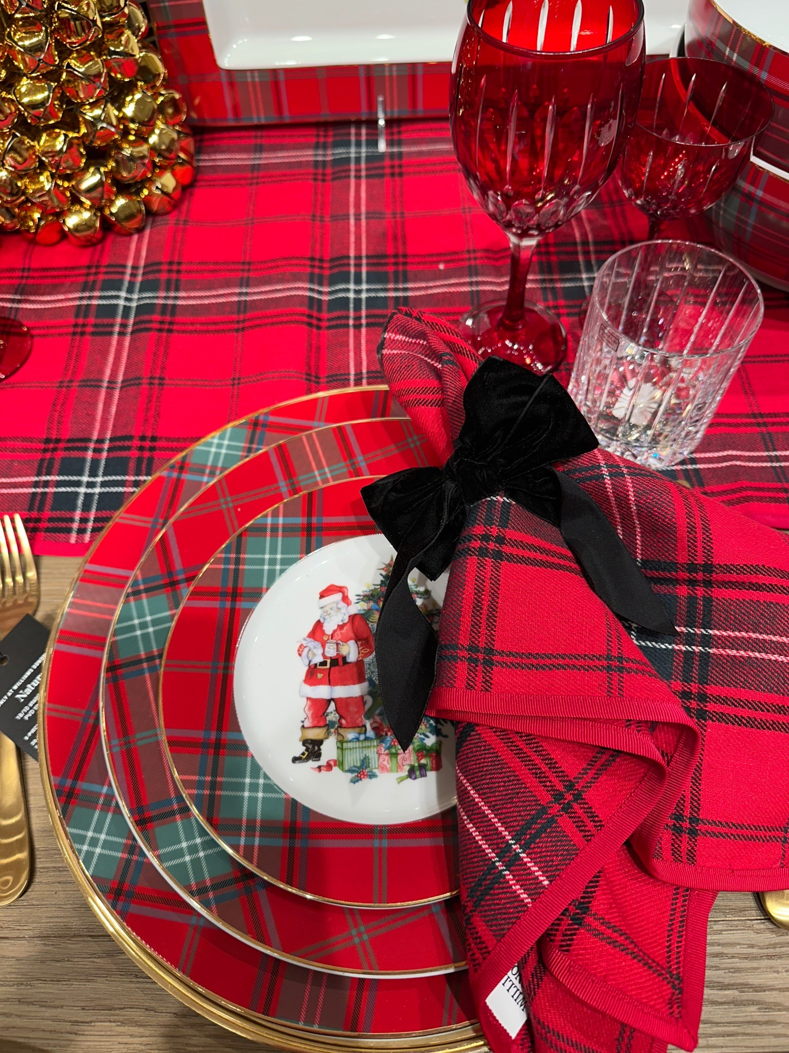 Classic Christmas dishes and table linens in red, green and black.  Traditional plaid plates, plaid tablecloth and Christmas napkins with black velvet bow.  Crystal glassware and gold flatware along with a gold bell tree.

#LTKSeasonal #LTKHoliday #LTKHome