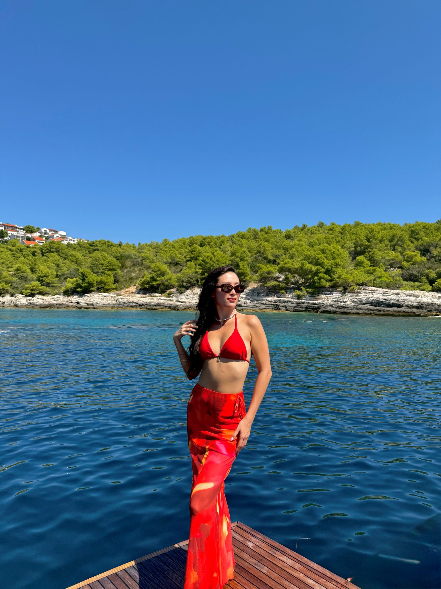 a red triangle bikini top with a gold cowrie shell pendant detail paired with a red and orange floral print bias-cut maxi sarong skirt, styled with tortoiseshell oval sunglasses and a layered shell pearl necklace for a yacht day on the croatian coast. the bold red color story and silky floral sarong make this the ultimate mediterranean boat day swim look. perfect for hvar yacht days, croatia sailing trips, dalmatian coast vacations, and mediterranean boat outings. shop this look linked above.
