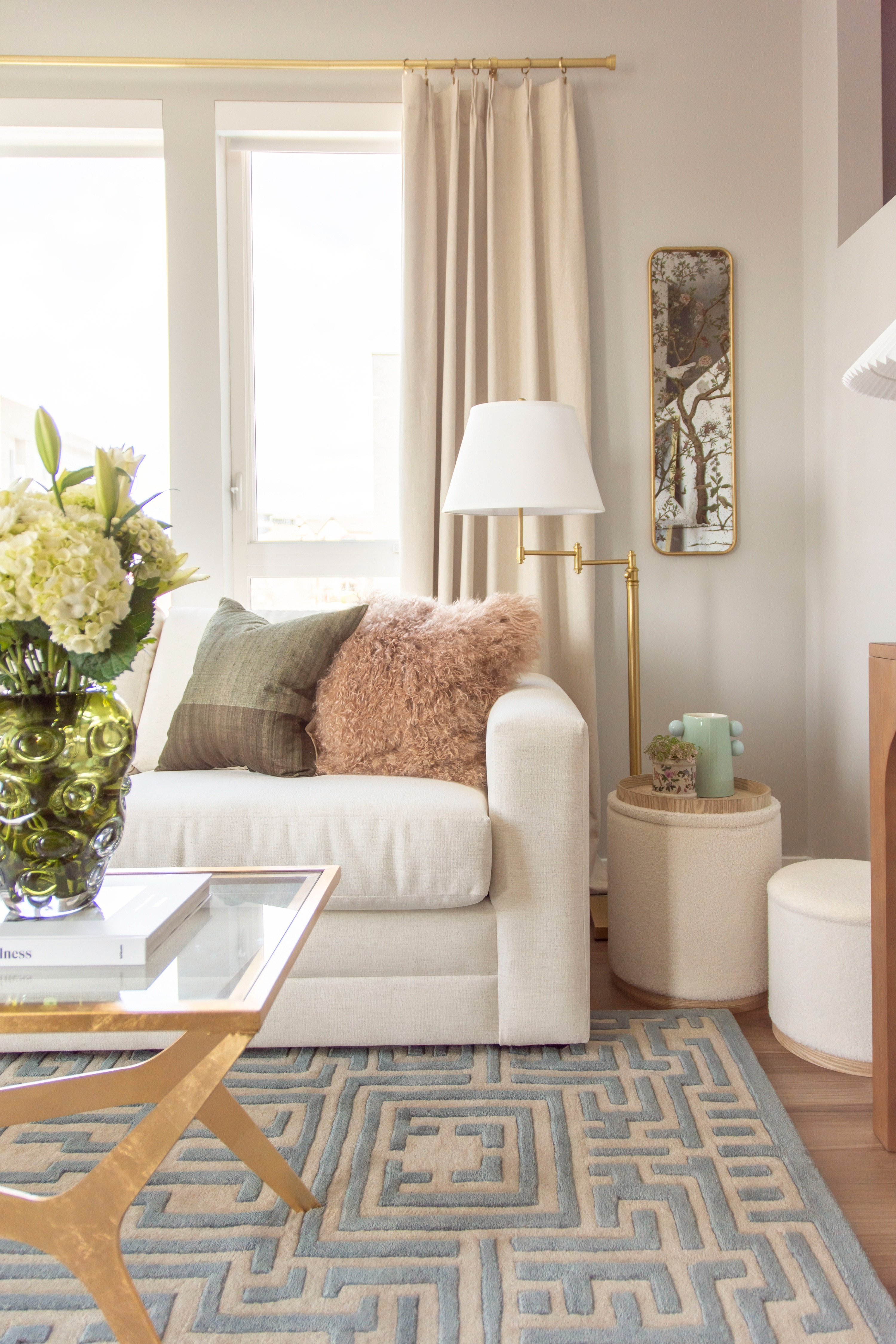 Neutral-toned living room with a cozy modern eclectic vibe. This stylish space features a boucle accent chair, a vintage floral armchair, and a textured green ottoman topped with a decorative tray and potted greenery. A sleek console table is styled with stacked coffee table books and small decor, while a wall-mounted TV blends into the design as framed art. Warm textures, layered textiles, and natural light complete this inviting home decor look, perfect for chic apartment living and timeless interior inspiration. 

 