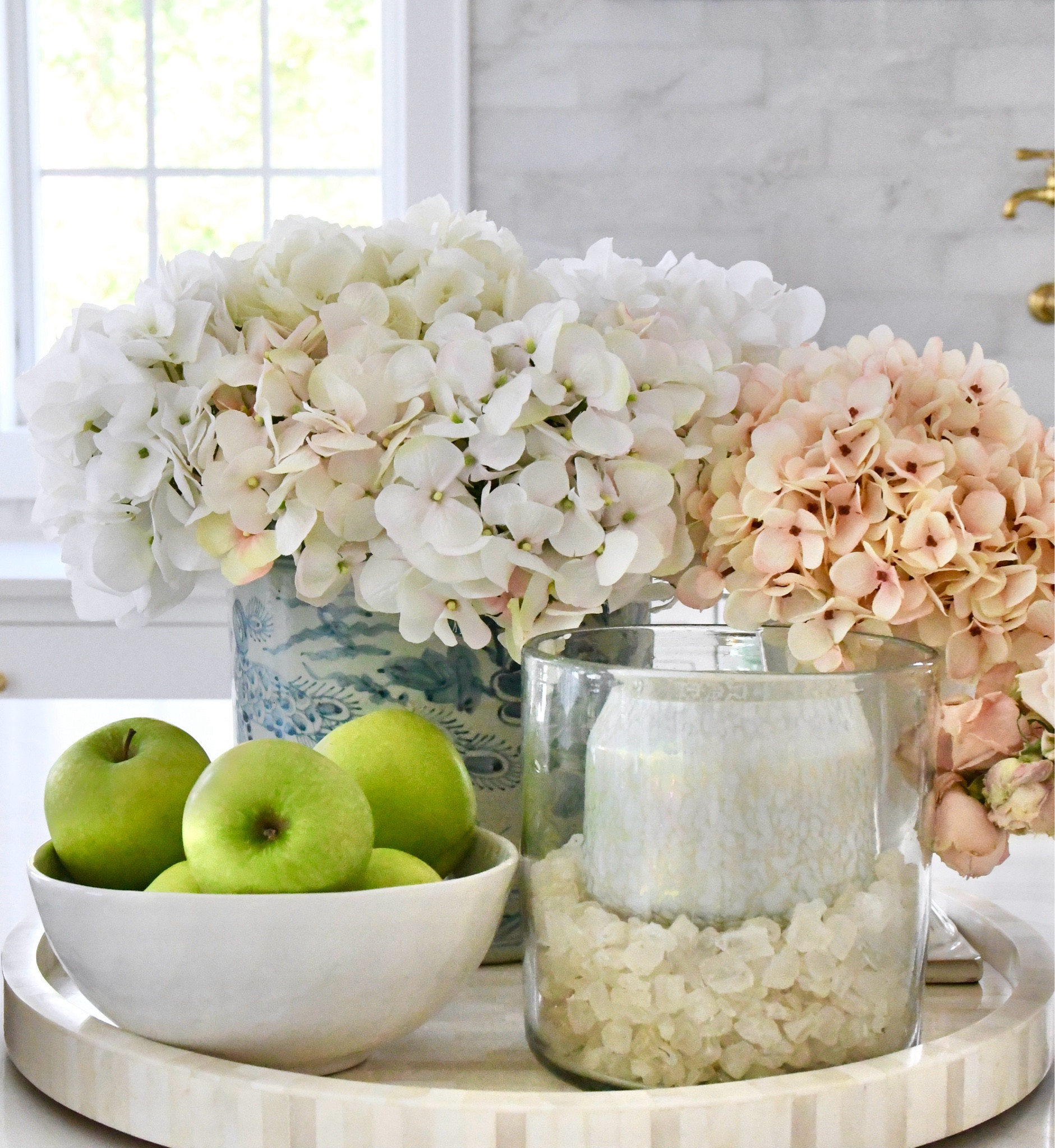 Simple kitchen styling tip:
Begin with a round tray. Add 1 planter filled with faux hydrangeas, 1 hurricane with glass rocks and a candle, and a bowl full of fruit. This is a great look for summer  You can switch your fruit and candle to fit the season! 

#LTKhome