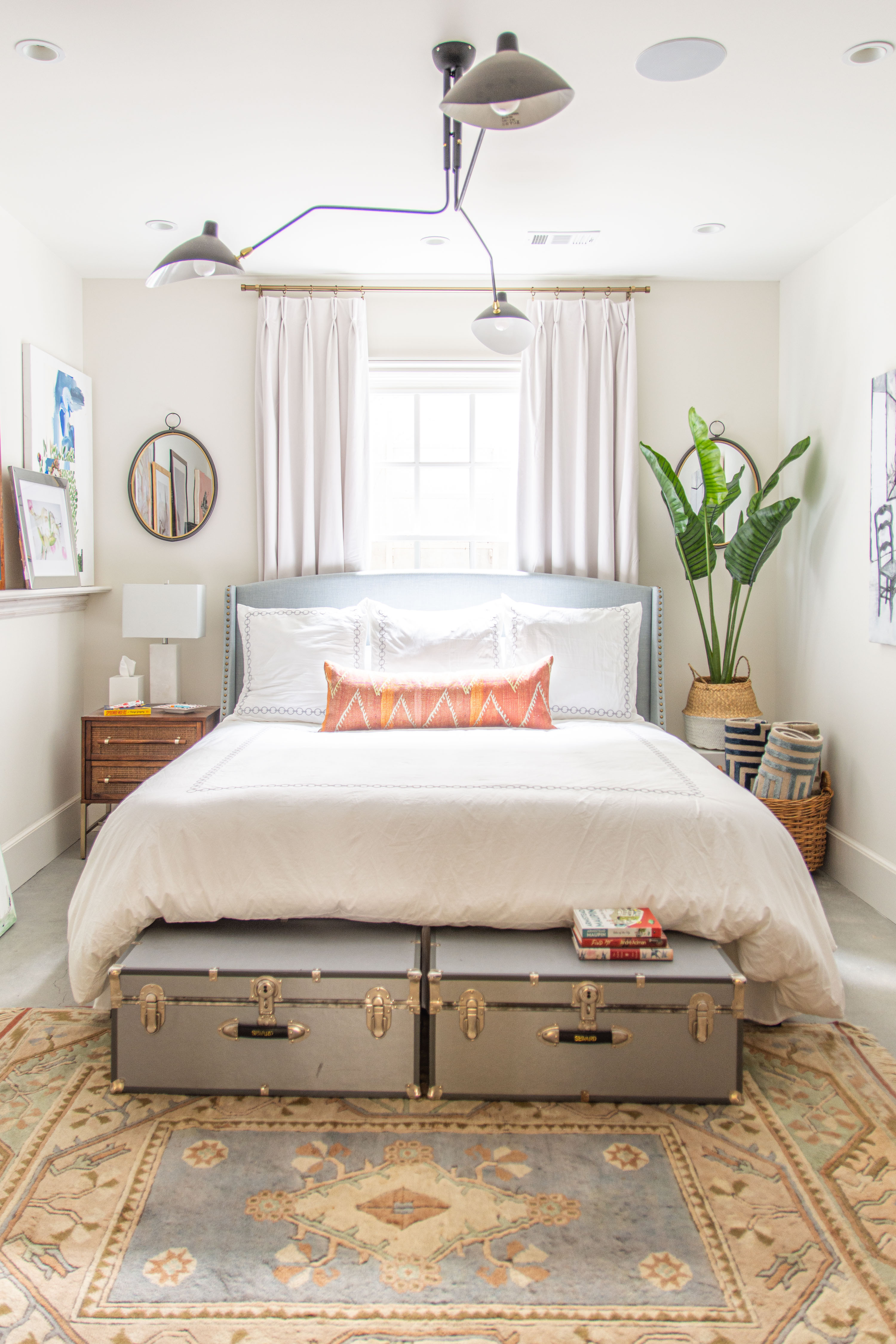 Bright and airy modern bedroom with a clean neutral palette. This space features a light upholstered bed frame with crisp white bedding, accented by a long lumbar pillow in warm tones. Matching nightstands with modern lamps add symmetry, while layered storage trunks at the foot of the bed double as functional decor. A vintage-inspired area rug grounds the room with soft patterns and muted colors. Tall curtains frame the window, enhancing natural light, while a woven basket with rolled blankets and a large potted plant add warmth and texture. The contemporary ceiling light fixture completes the modern yet cozy design. 

 