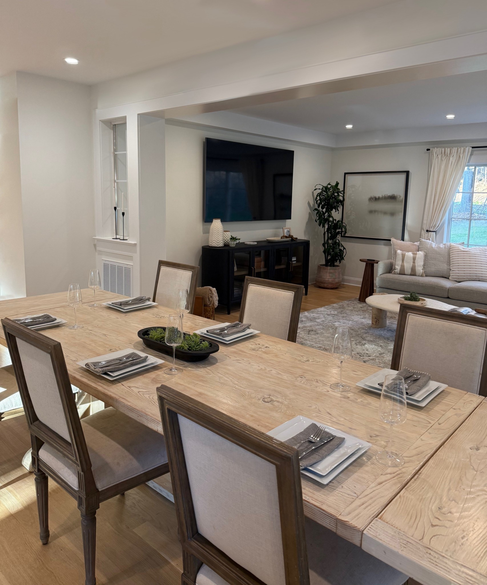 A stylish dining room scene seamlessly flows into the adjacent living area, where a sleek console and mounted TV provide a modern focal point. The dining space is anchored by a warm, inviting table surrounded by thoughtfully chosen seating, creating the perfect blend of comfort and sophistication. Soft lighting and a neutral palette tie both areas together, offering a cohesive and welcoming open-concept layout. 