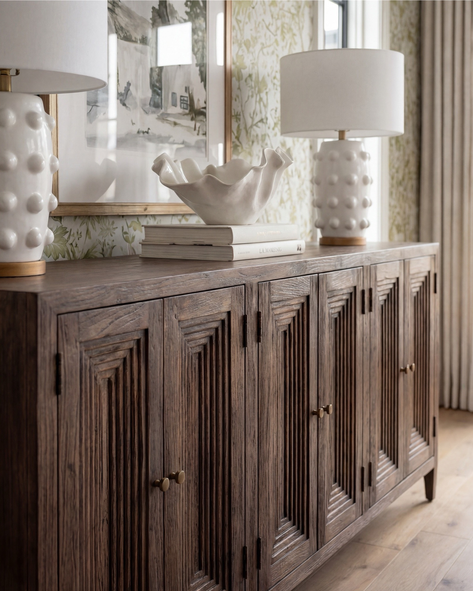 There’s something about a light filled, nature driven dining room that feels effortlessly welcoming the moment you step inside. This space blends soft pattern, warm wood, and organic lines in a way that feels both elevated and incredibly livable.

The pedestal dining table brings so much presence with its sculpted base and natural wood grain. I paired it with clean, curved dining chairs in a soft ivory upholstery to keep the look airy and balanced. On the sideboard, the deeper wood tone adds richness and grounds the entire room.

Styling is all about quiet impact here — a large ceramic vase filled with fresh greenery, a simple tray, and layered candles that keep the table looking intentional without taking away from the architecture around it. The twin lamps on the sideboard add symmetry and that perfect soft glow.

And that wallpaper — the muted green florals feel traditional in the best way, adding depth and texture without overwhelming the room. The dark, vintage inspired rug anchors the whole palette, pulling in those moody greens and creating contrast against the lighter woods.

✨ Shop the Look
* Pedestal dining table
* Curved wood dining chairs
* Green floral wallpaper
* Dark vintage inspired rug
* Ceramic statement vase
* Gold rim tray
* Sideboard table lamps

#LTKsale #LTKhome #LTKstyletip