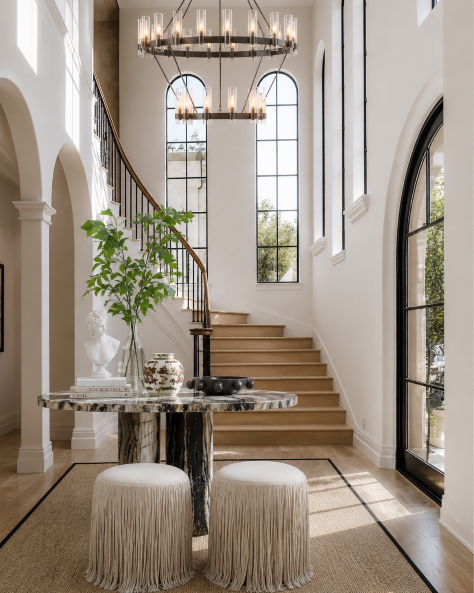 A few simple pieces can make an entryway feel so much more finished. A round table gives you a natural focal point, the stools add extra texture without taking up visual space, and the oversized plant helps balance all the height from the stairs, windows, and chandelier.

It’s a good formula for a large entry that feels empty: one substantial table, layered decor, something sculptural, and a little softness underfoot.

This post contains affiliate links, and I may earn a small commission from purchases at no additional cost to you.